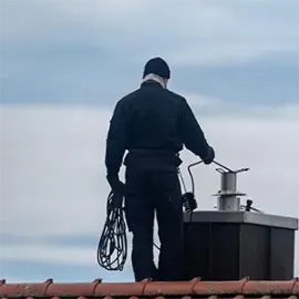 Person in dark work clothes stands on a rooftop adjusting a metal chimney under a cloudy sky