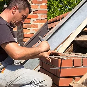 Worker laying bricks on a rooftop next to a chimney, using a trowel and level.
