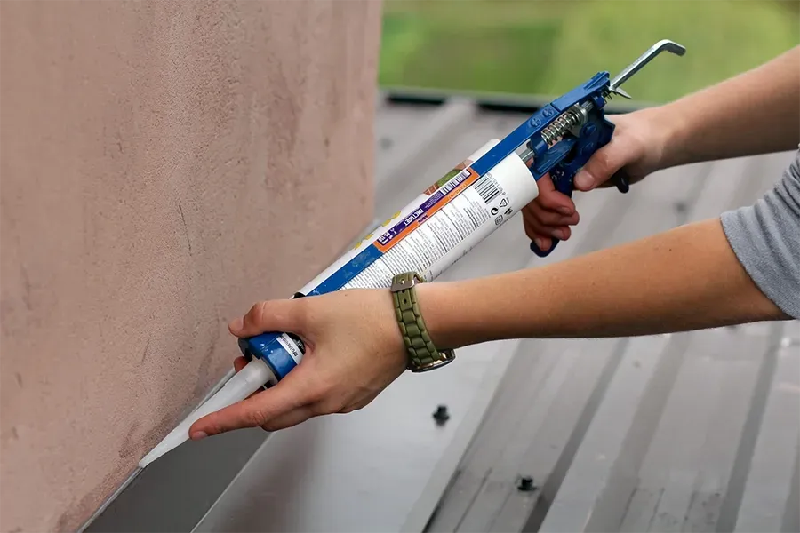 Hands applying sealant with a caulking gun along a seam on a roof edge.
