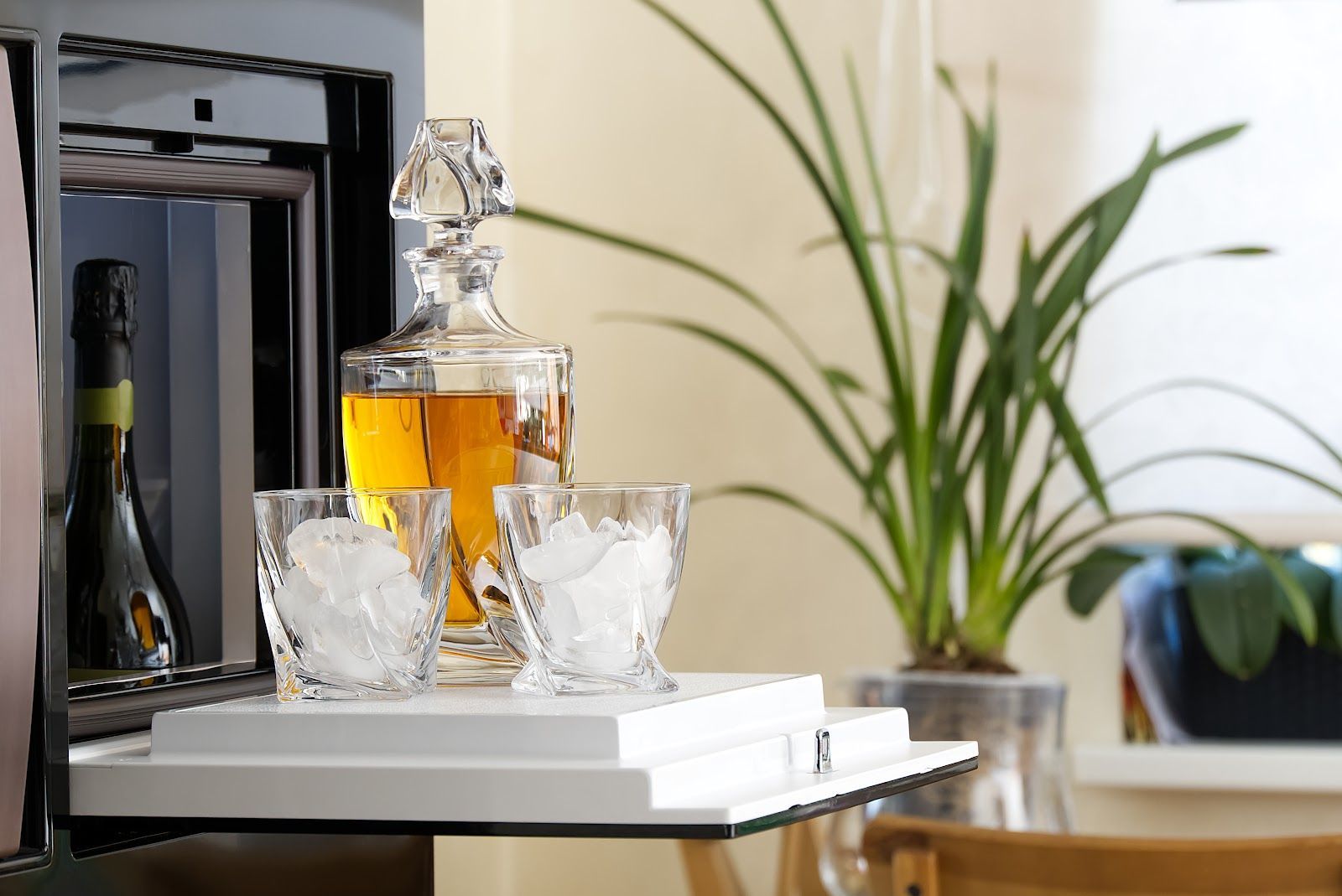 A home bar setup: decanter, glasses with ice, and a plant near a mini fridge.