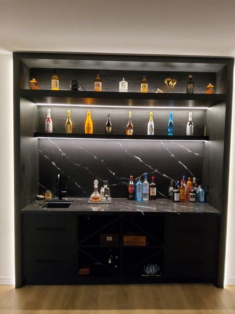 Dark home bar with backlit shelves displaying liquor bottles, a sink, and cabinets.