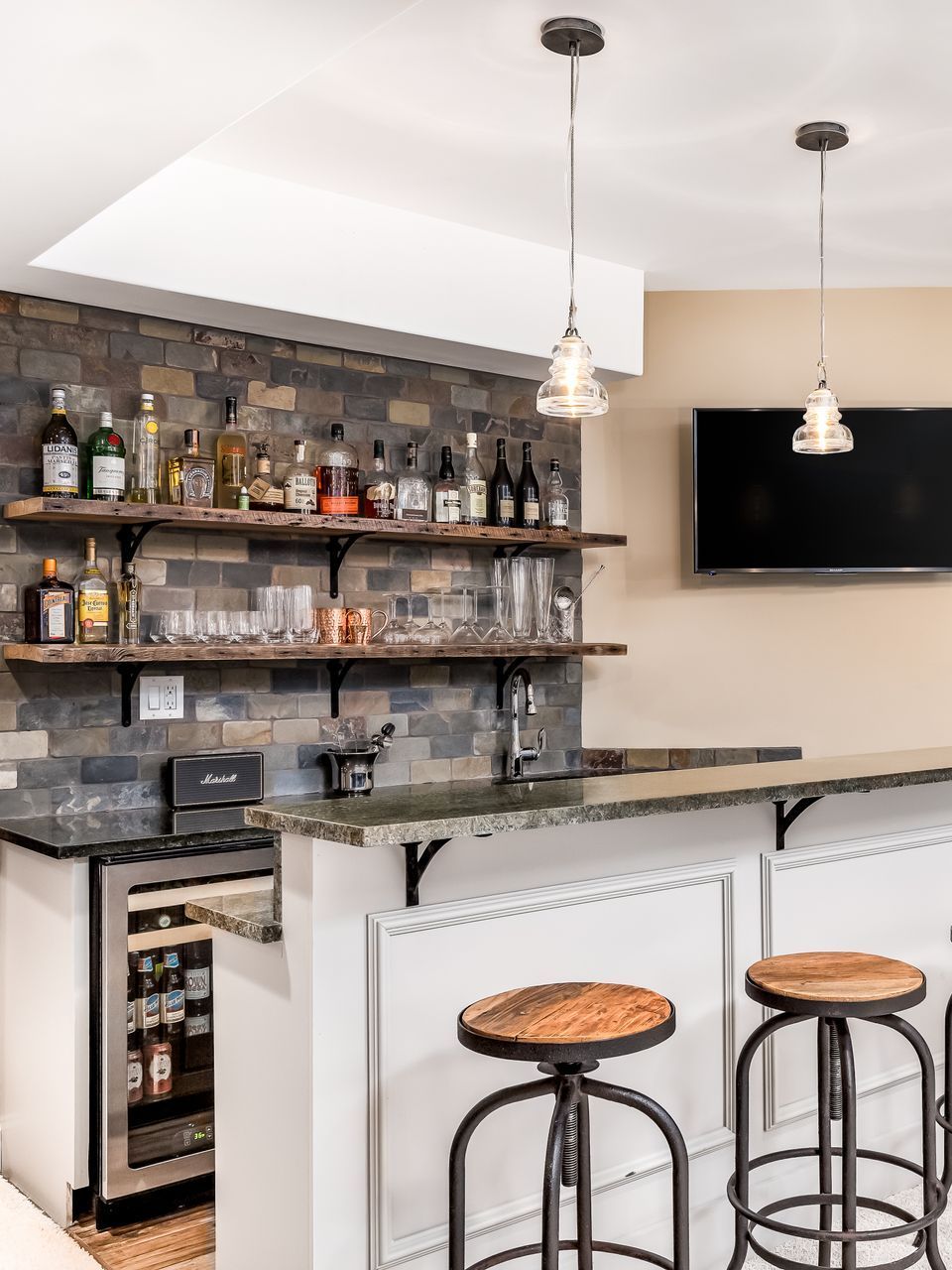 Cozy home bar with stone accent wall, floating shelves of liquor, countertop, and two bar stools. A mini fridge is on the left.