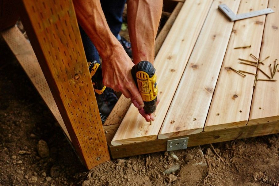 A person uses a power drill to fasten wooden deck boards to a frame outdoors.