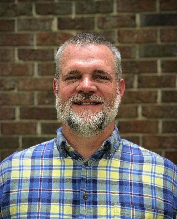 A man with a beard and a plaid shirt is smiling in front of a brick wall.