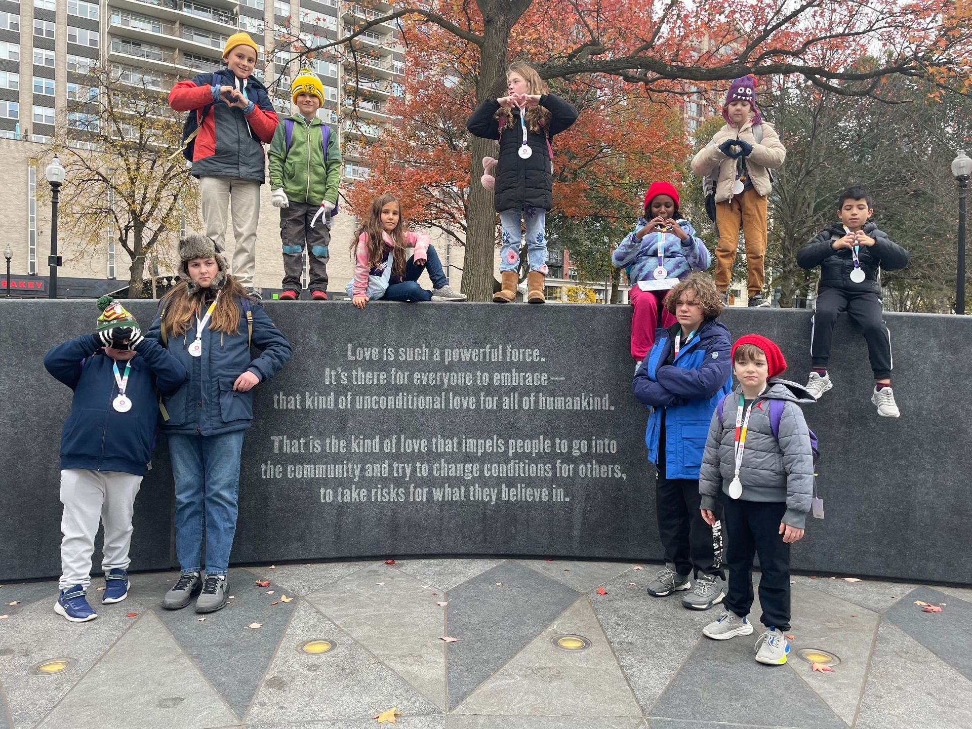 Children posing on and around a monument in a park, with autumn trees and buildings behind.