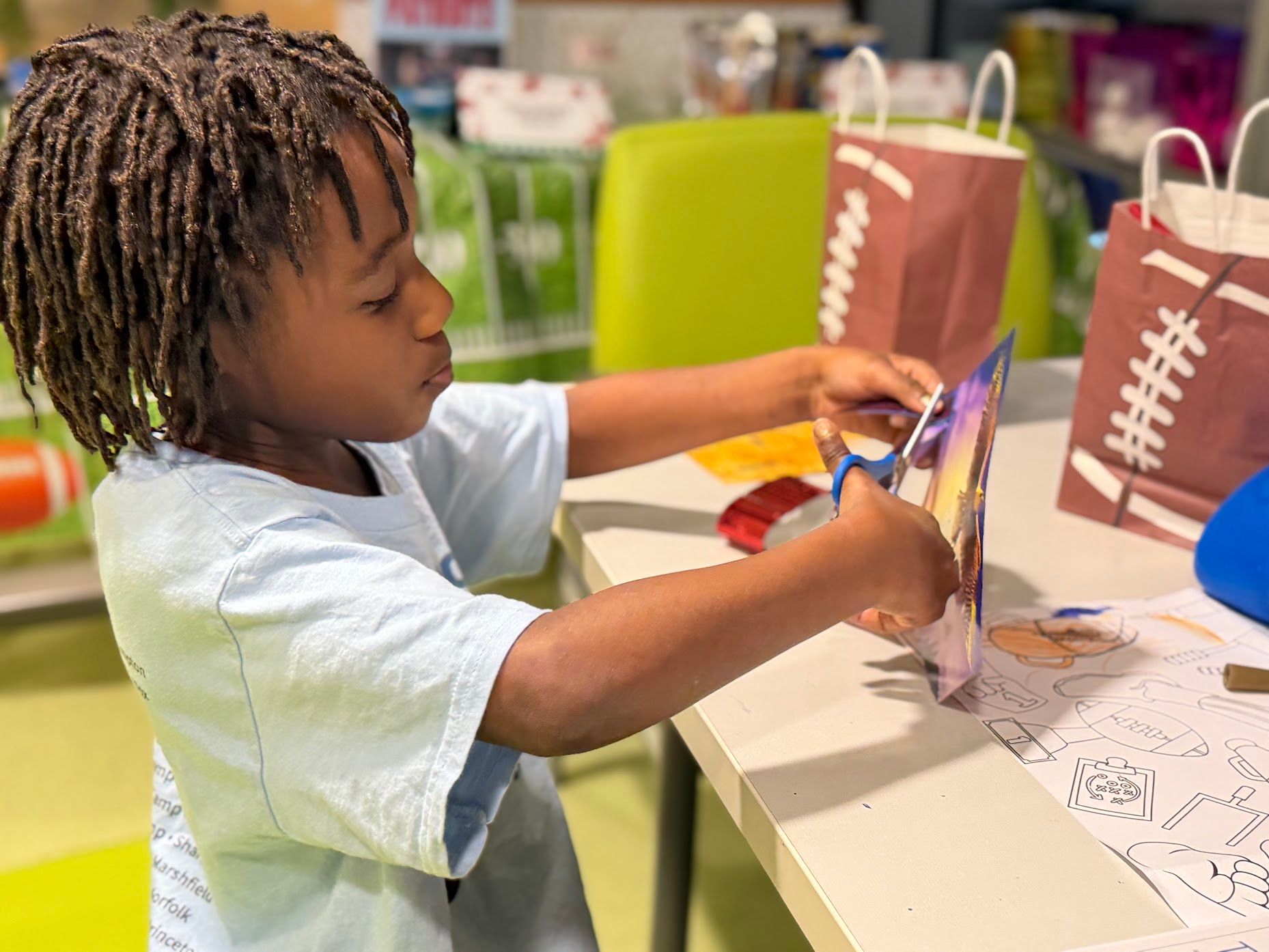 Child cutting paper at a classroom table with scissors, surrounded by art supplies and coloring pages