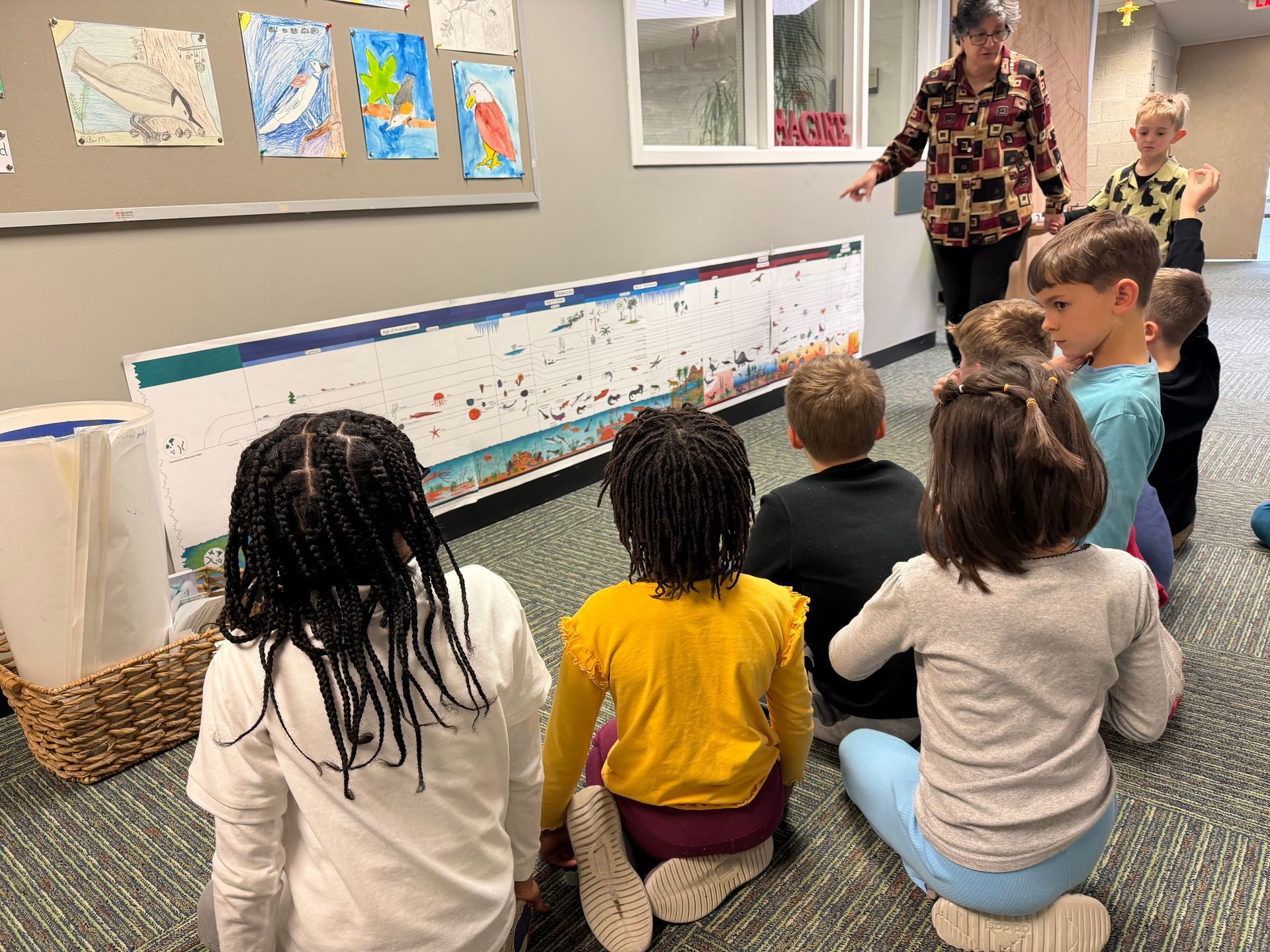 Children kneel in a classroom while a teacher points to the timeline of life on the wall.