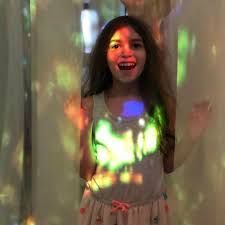 Smiling woman standing between curtains, lit by colorful projected lights.