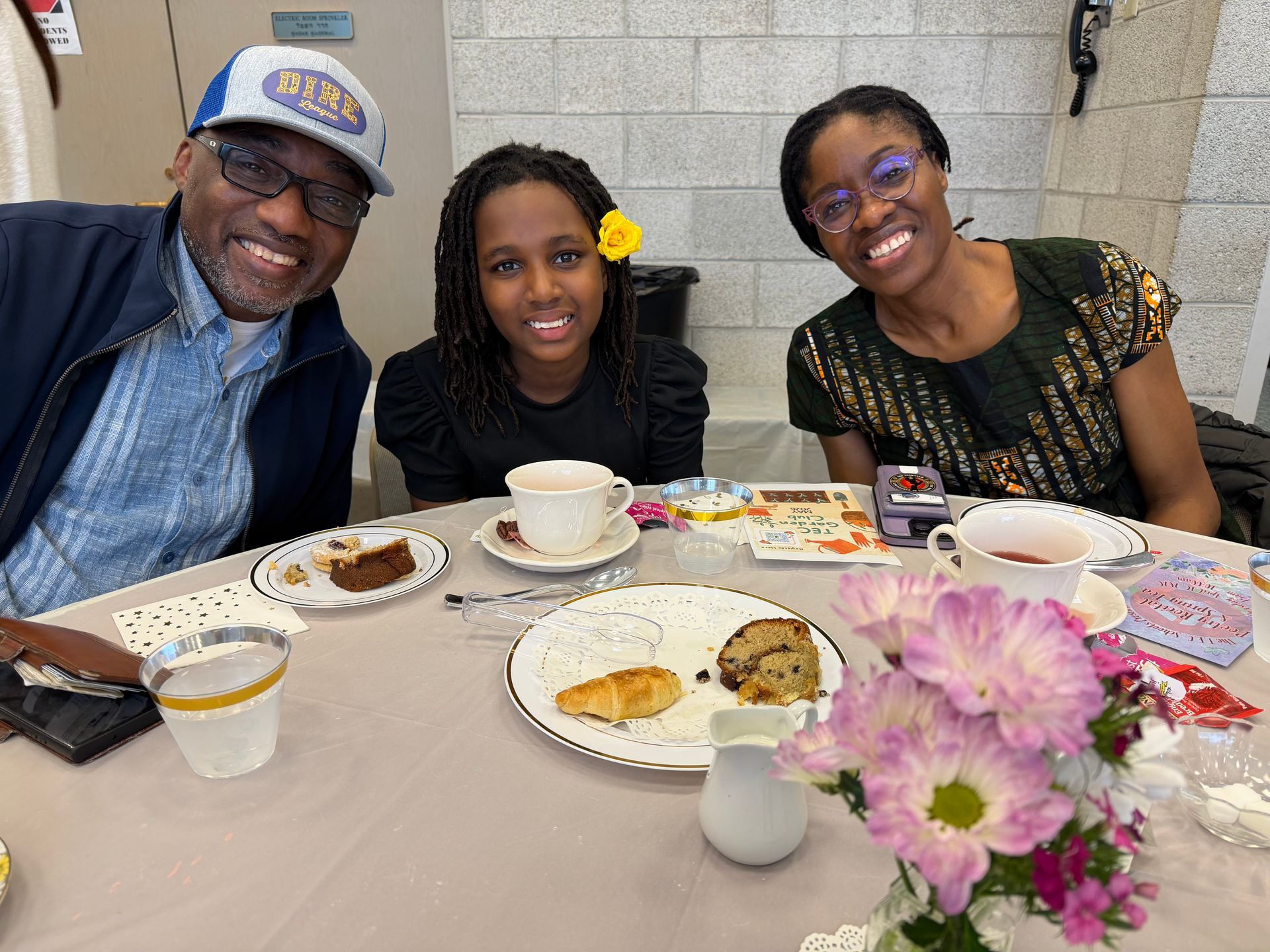 Three people smiling at a table with coffee, pastries, and flowers in a bright café