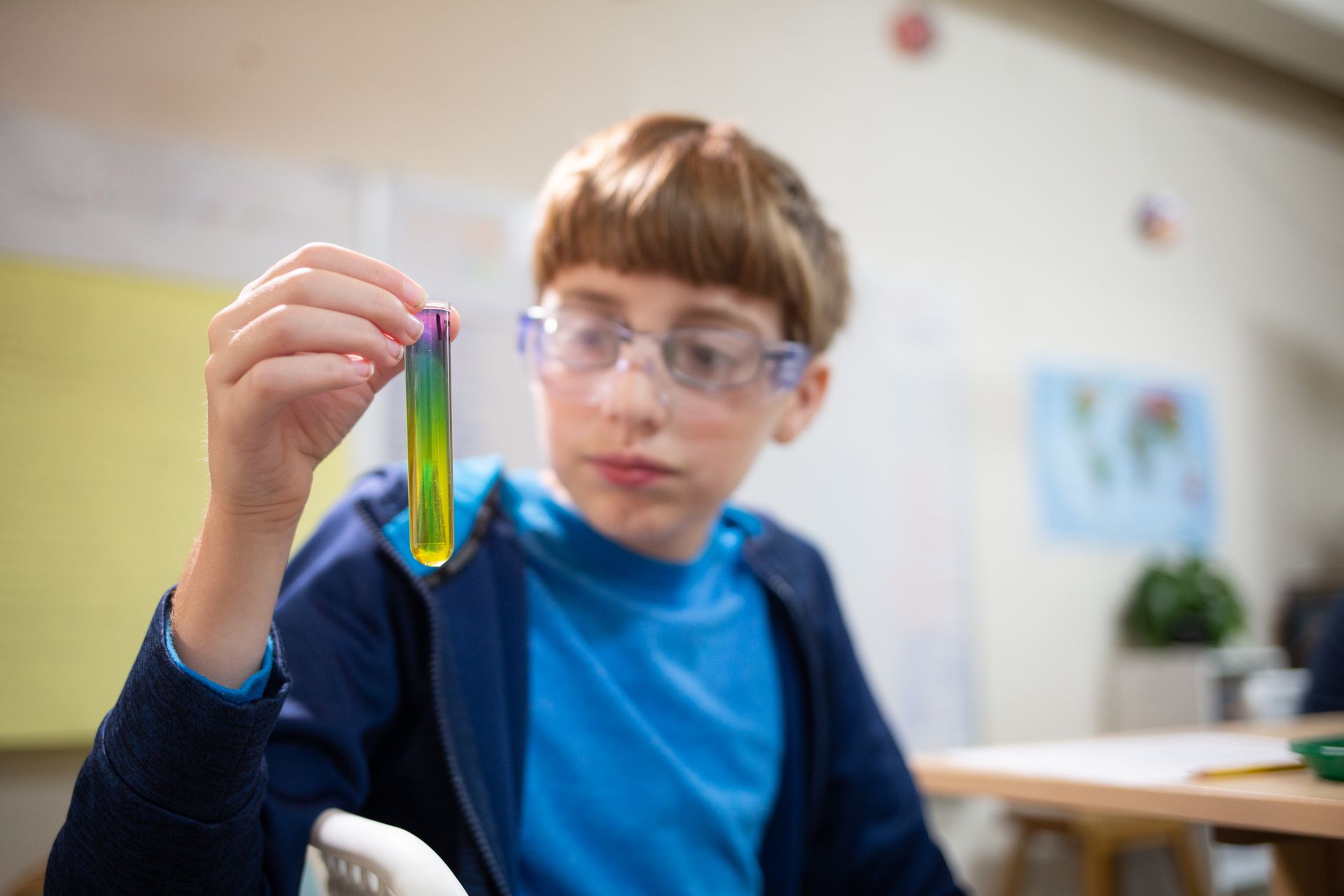 Child in a classroom holding a test tube with colorful liquid, wearing safety goggles.