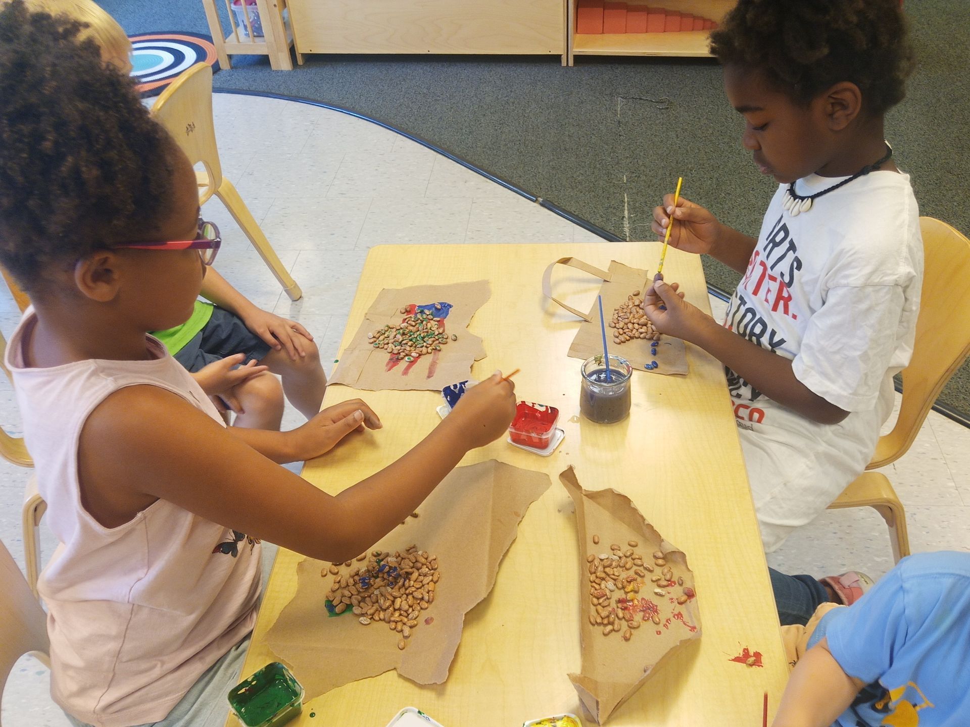 Children making arts and crafts at a table with beads, paper bags, and glue in a classroom