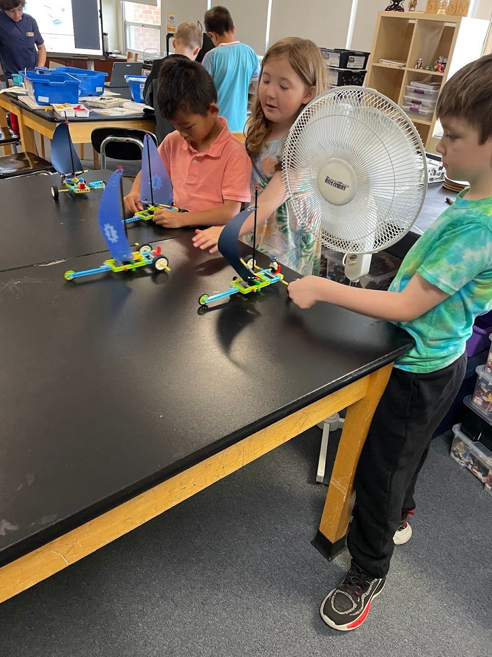 Children building with colorful LEGO at a classroom table, with a desk fan nearby.