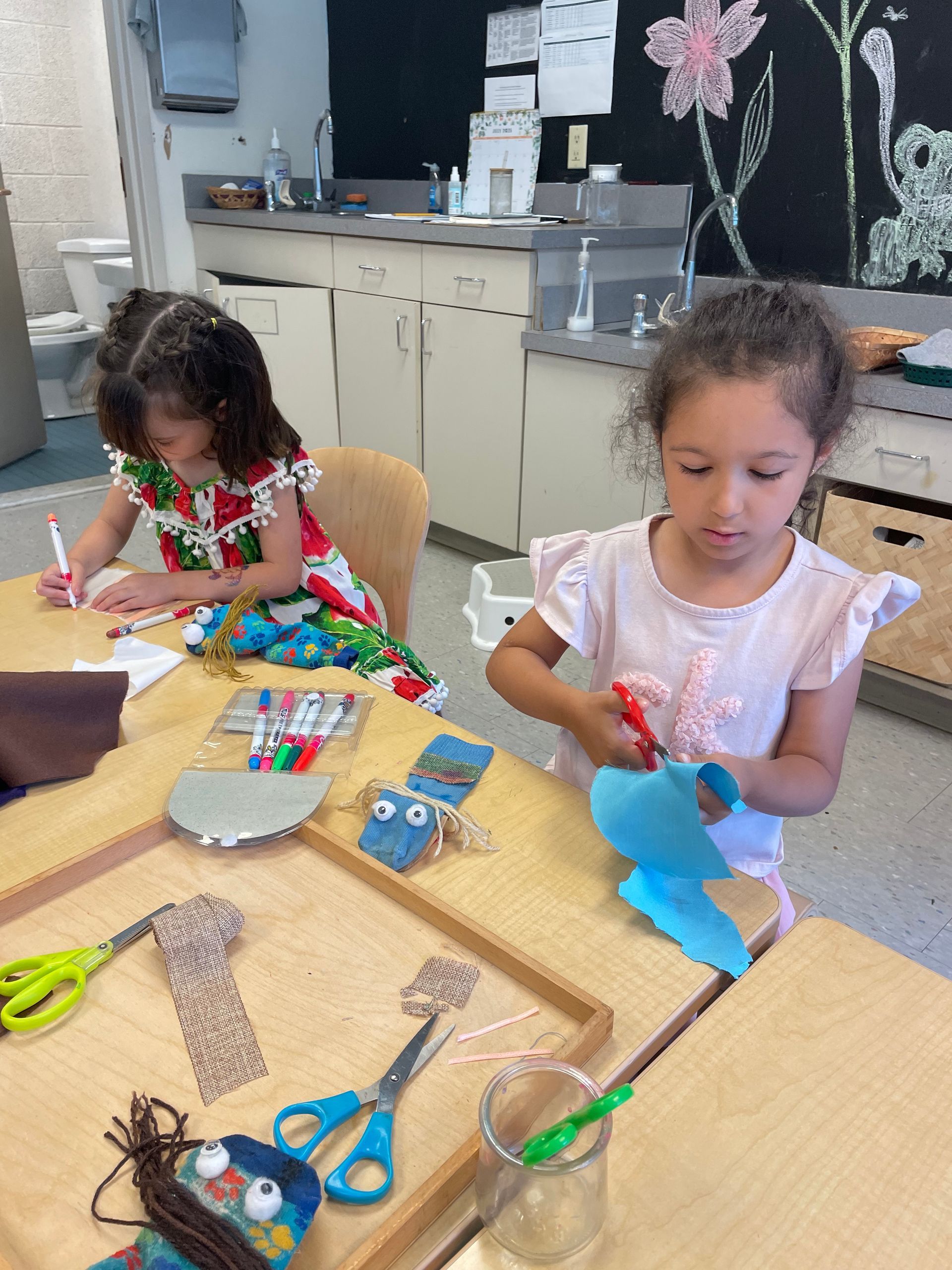 Two children crafting at a table with colorful paper, scissors, and glue in a classroom.