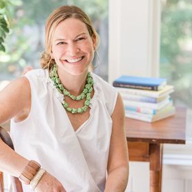 Smiling woman in a white blouse and green necklace sitting at a table with stacked books behind her