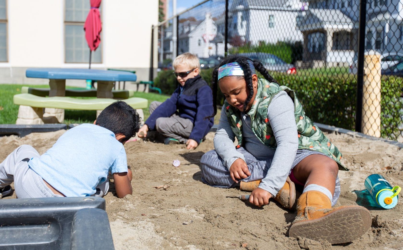 Children planting in a garden bed outdoors, with tools and a fence in the background