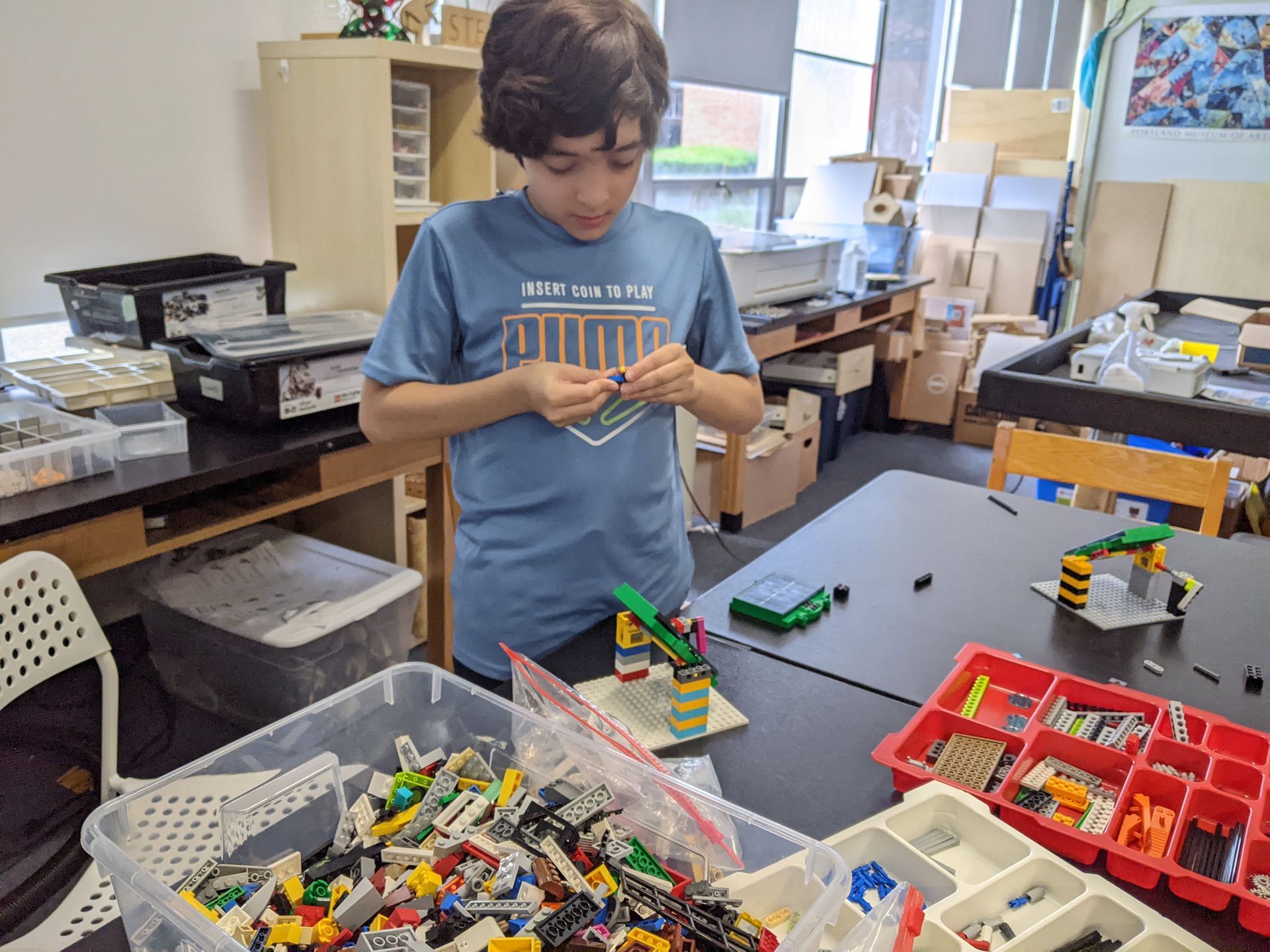 Child building with LEGO bricks at a classroom table, surrounded by colorful bins of pieces.