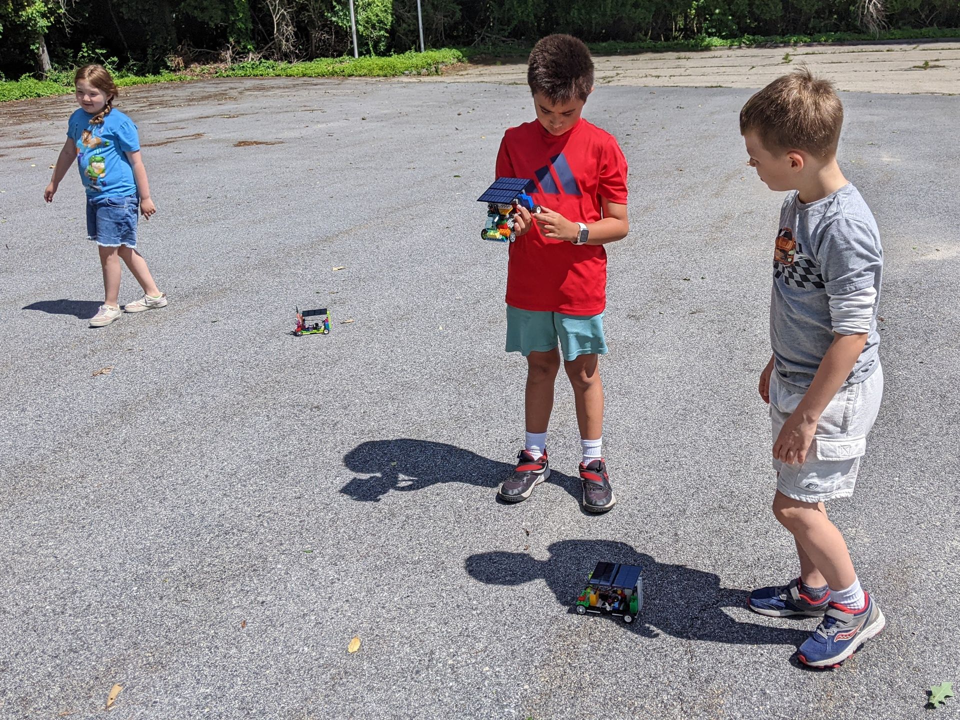 Three children play with toy cars on a sunlit paved road near trees.