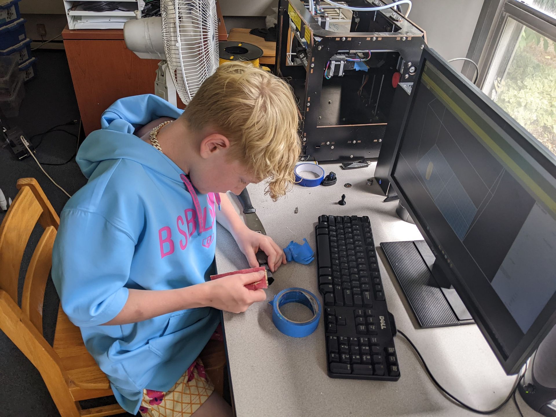 Person in a blue hoodie working with tape beside a computer monitor and keyboard on a desk.