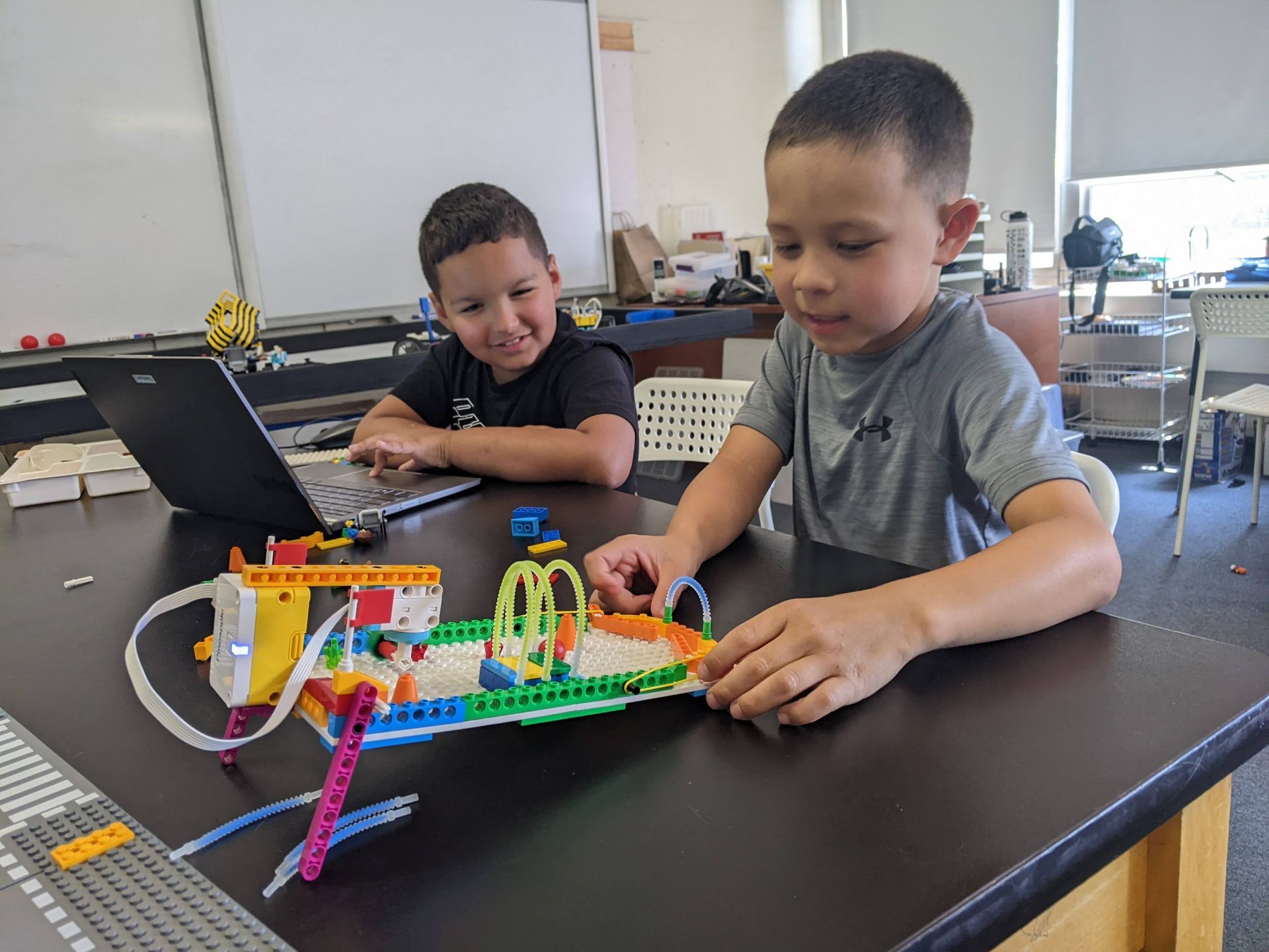Two children building with colorful LEGO bricks at a classroom table, with a laptop nearby.