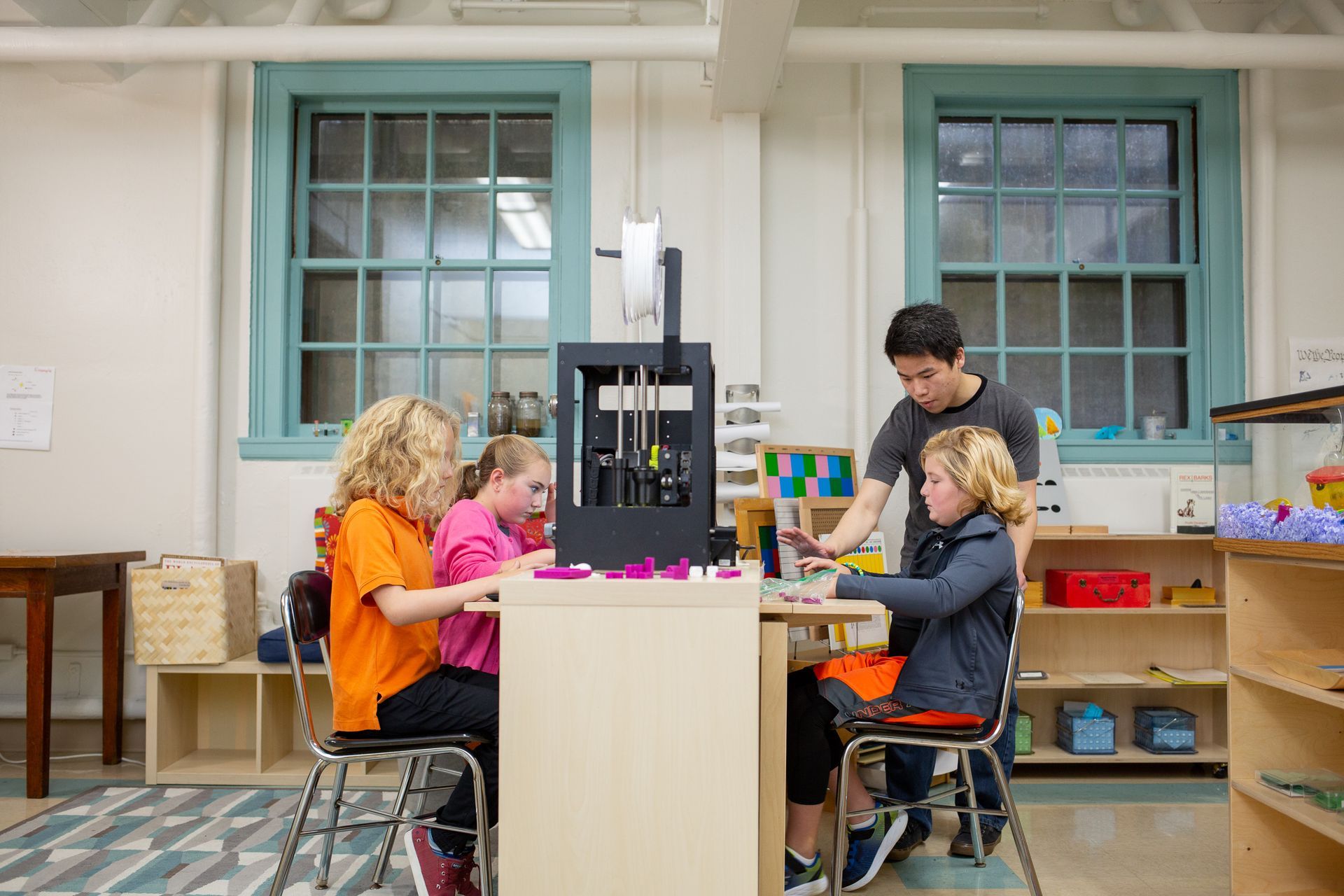 People gathered around a table in a bright classroom, working with a 3D printer and laptop
