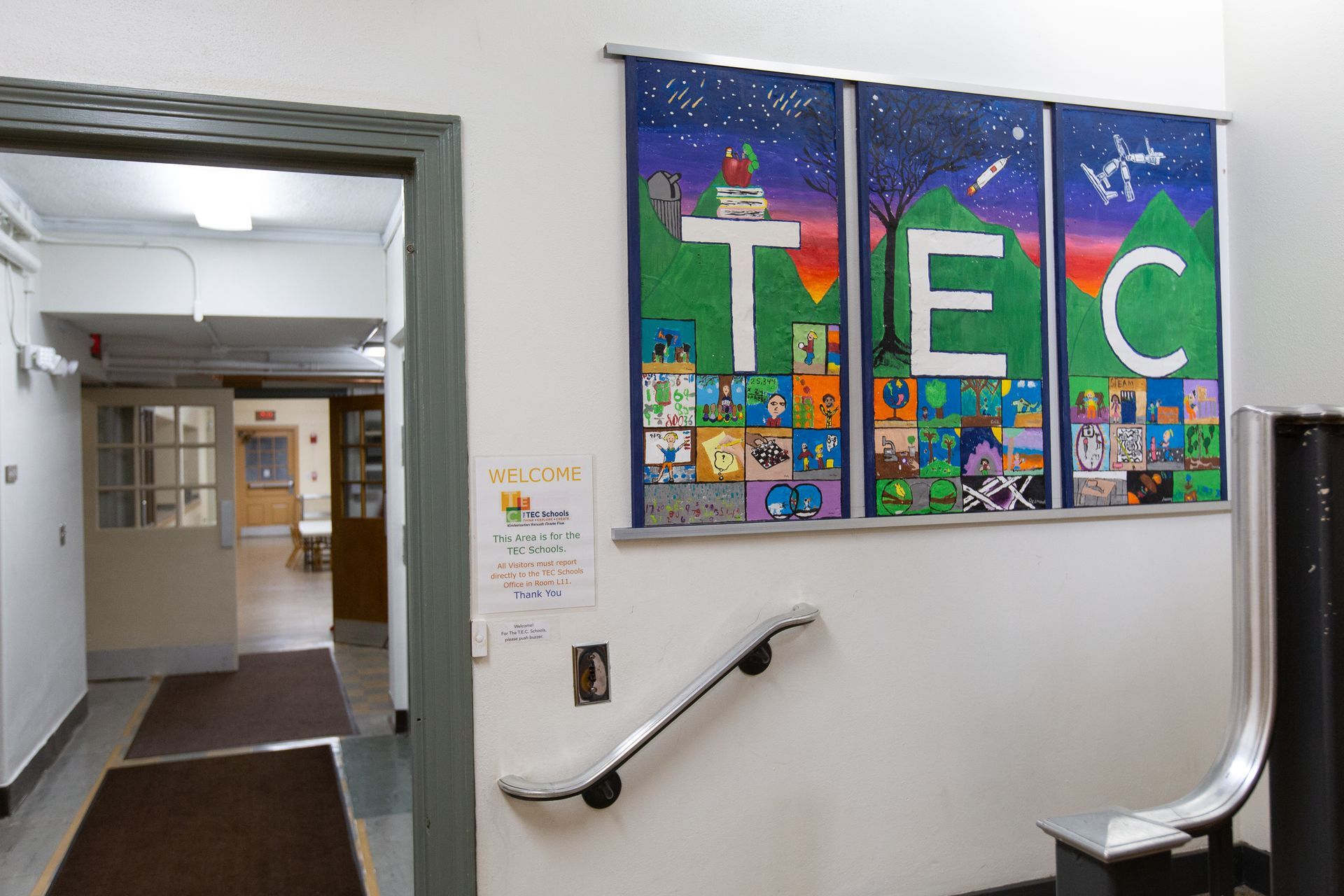 School hallway with colorful T E C wall murals and a stair handrail by an open doorway