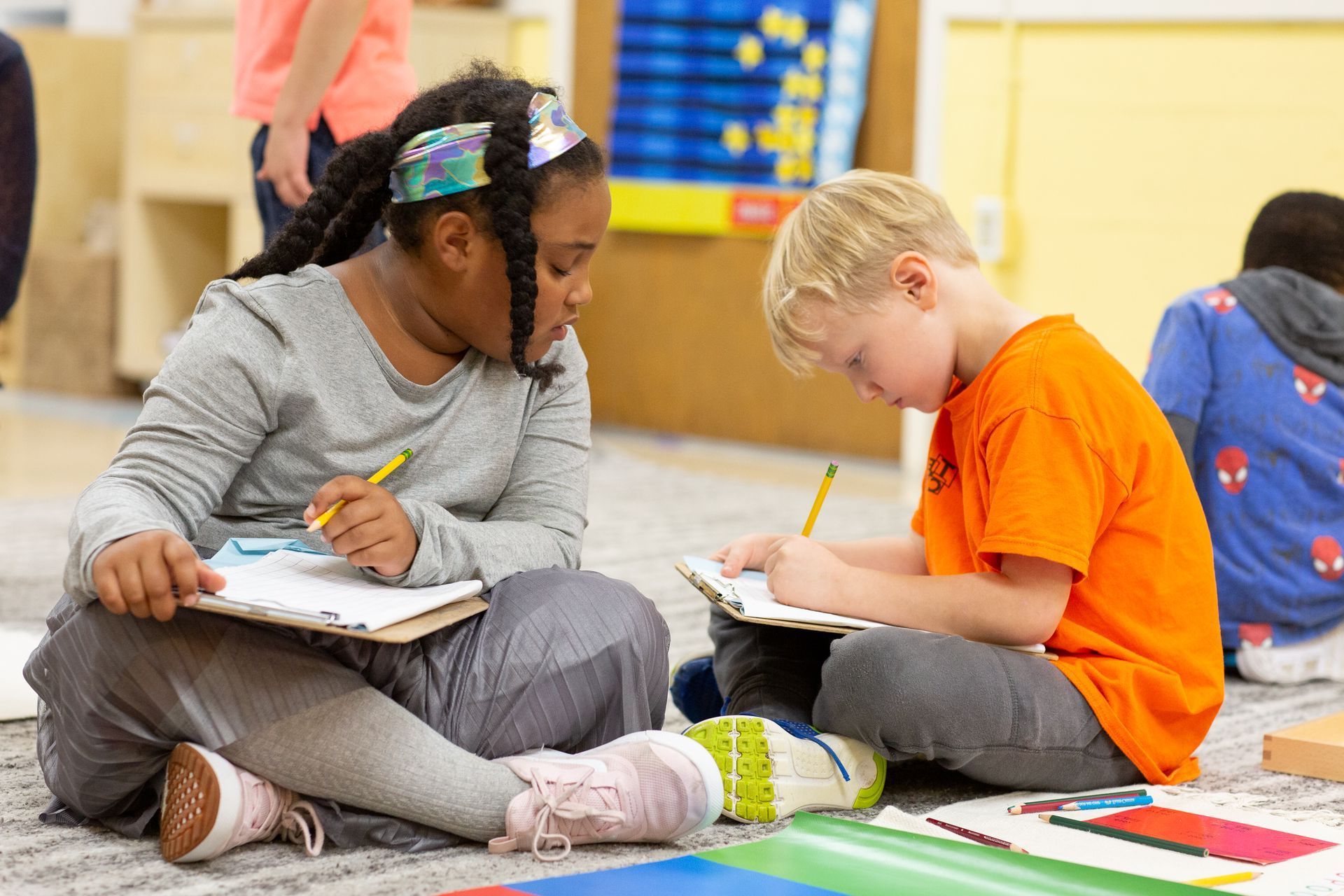 Two children sit on the floor, writing in notebooks during a classroom activity.