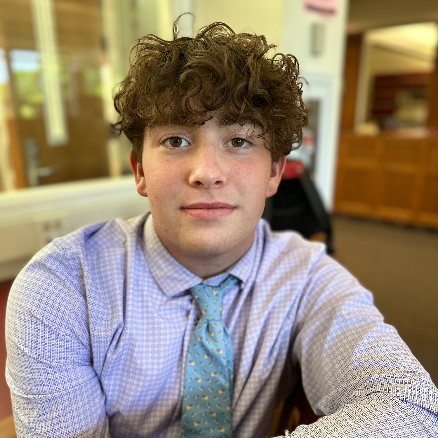 Young person in a light blue shirt and tie, seated indoors and looking at the camera.