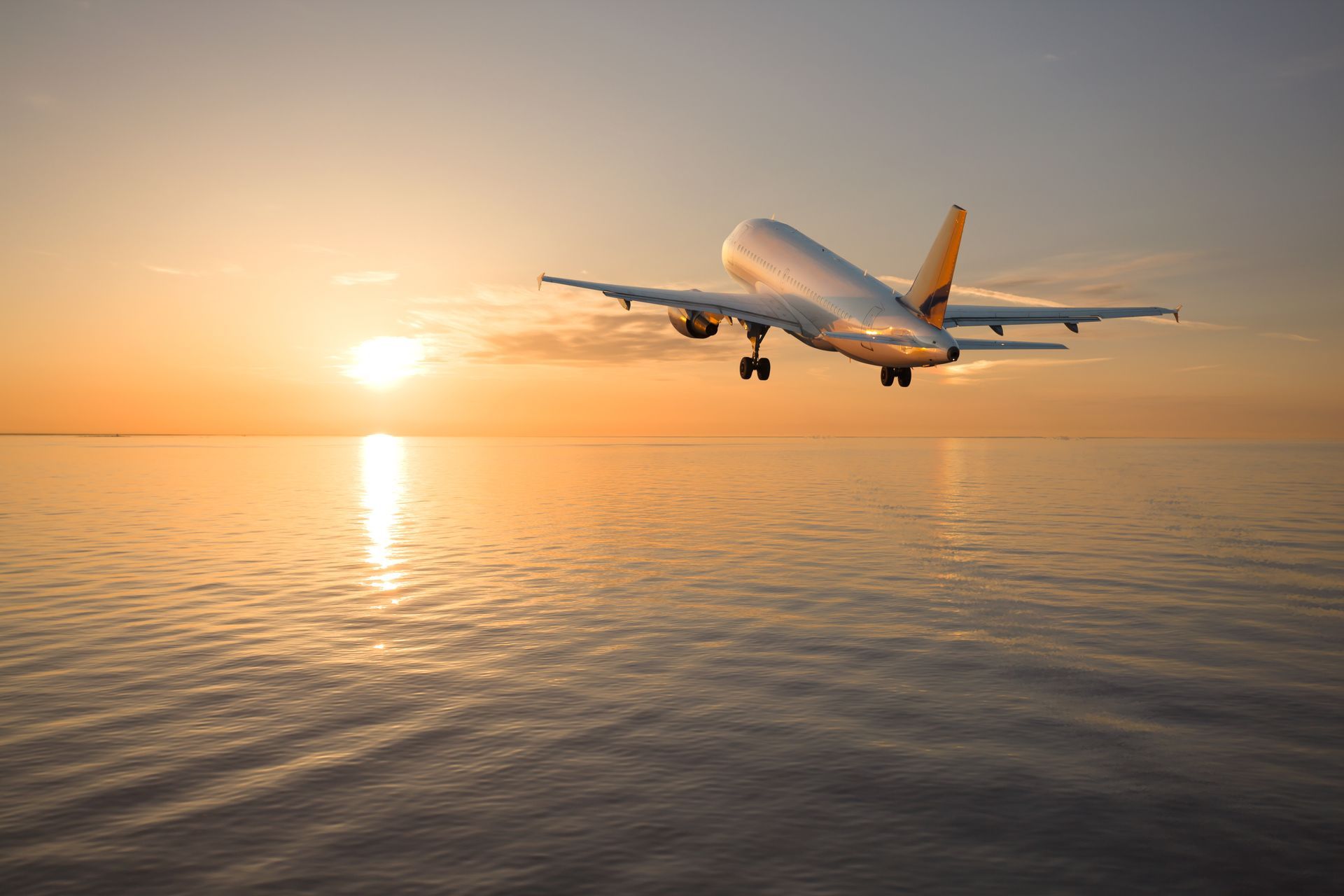 Airplane taking off over calm water at sunset, with orange sky and reflected light