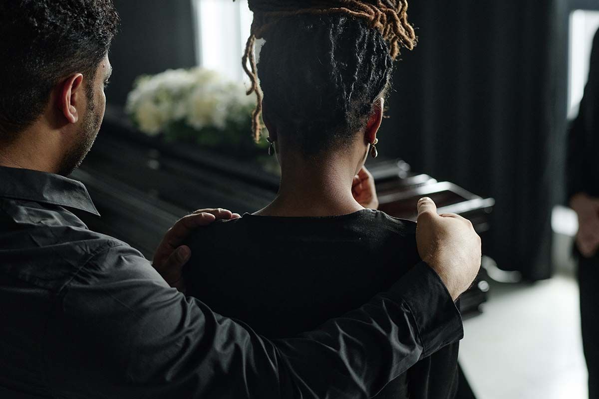 Two people sit beside a closed black casket in a dim funeral home