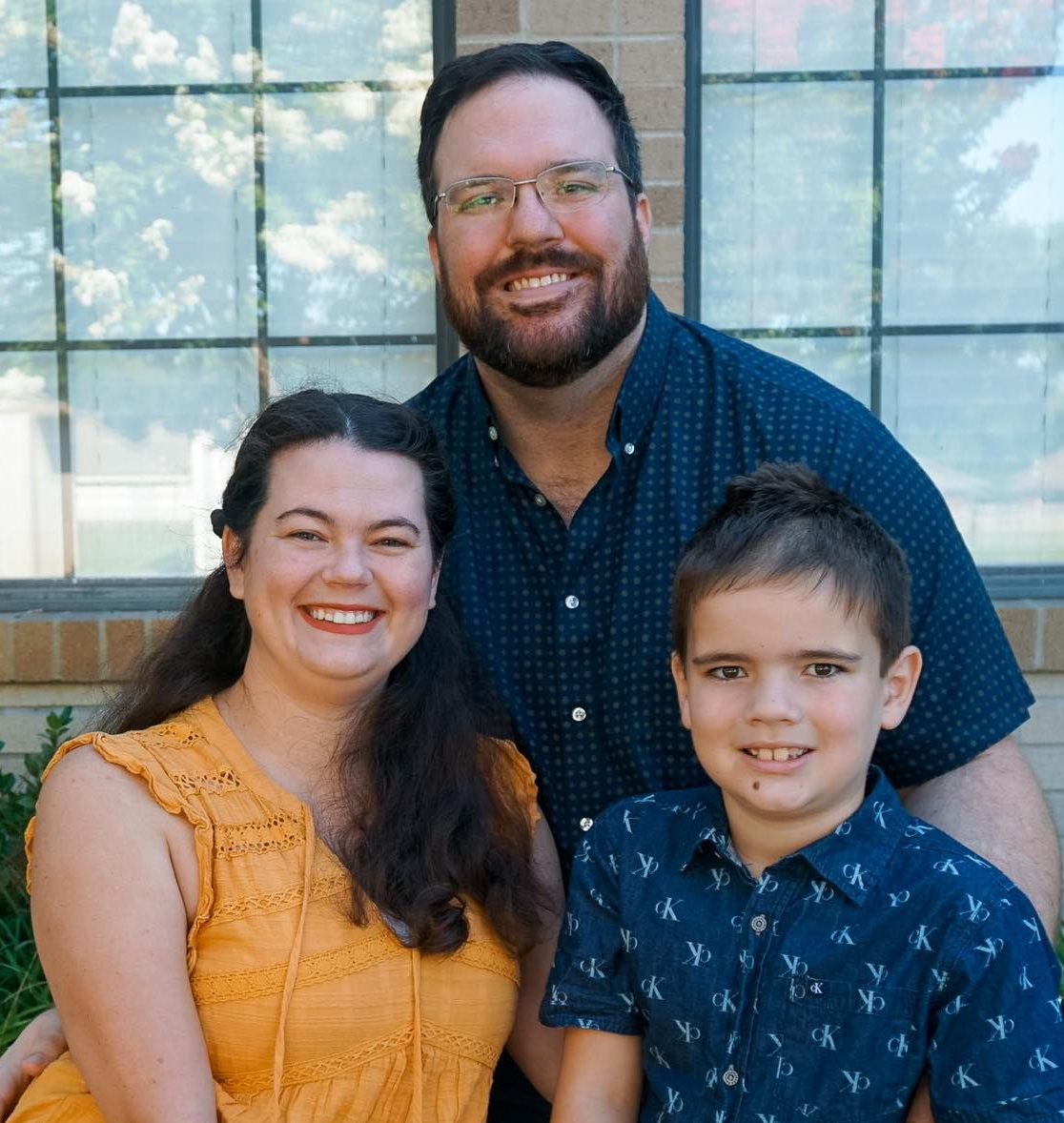 Family portrait: smiling woman in yellow top, man in blue shirt, and child in blue shirt. Outdoors.