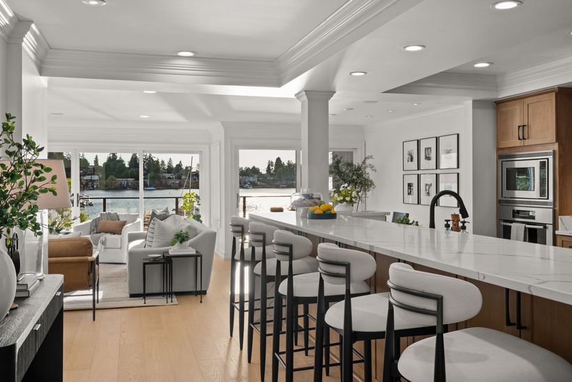 Bright open-concept kitchen and living room with a long island, barstools, and large windows overlooking trees.