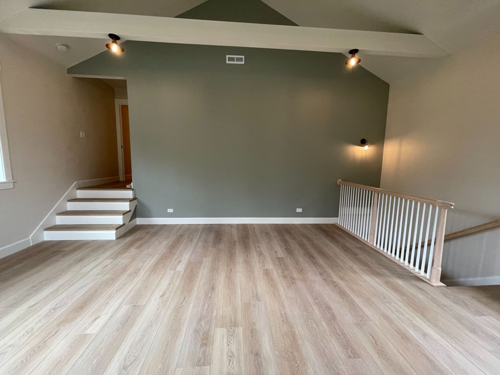 Empty living room with light wood floors, gray accent wall, staircase, and white railing.