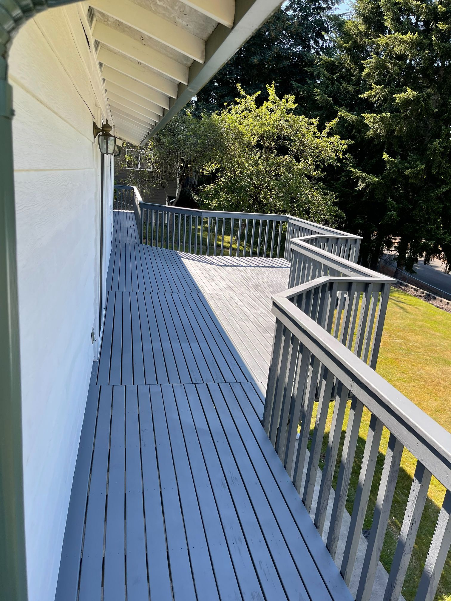 Grey wooden deck extending from a white house, with a green lawn and trees in the background.