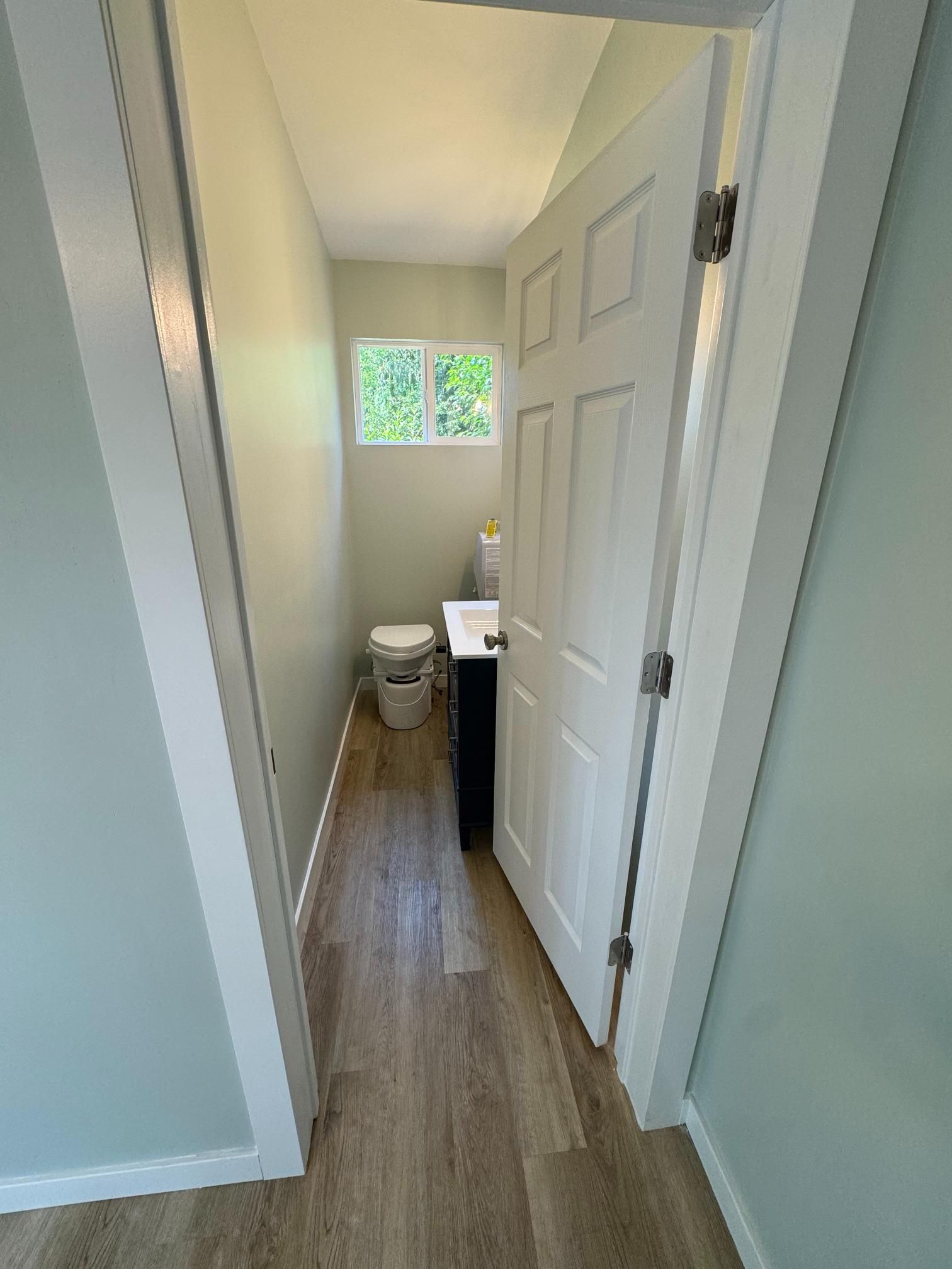Narrow bathroom with white door, toilet, sink, and window. Light blue walls, light brown floor.