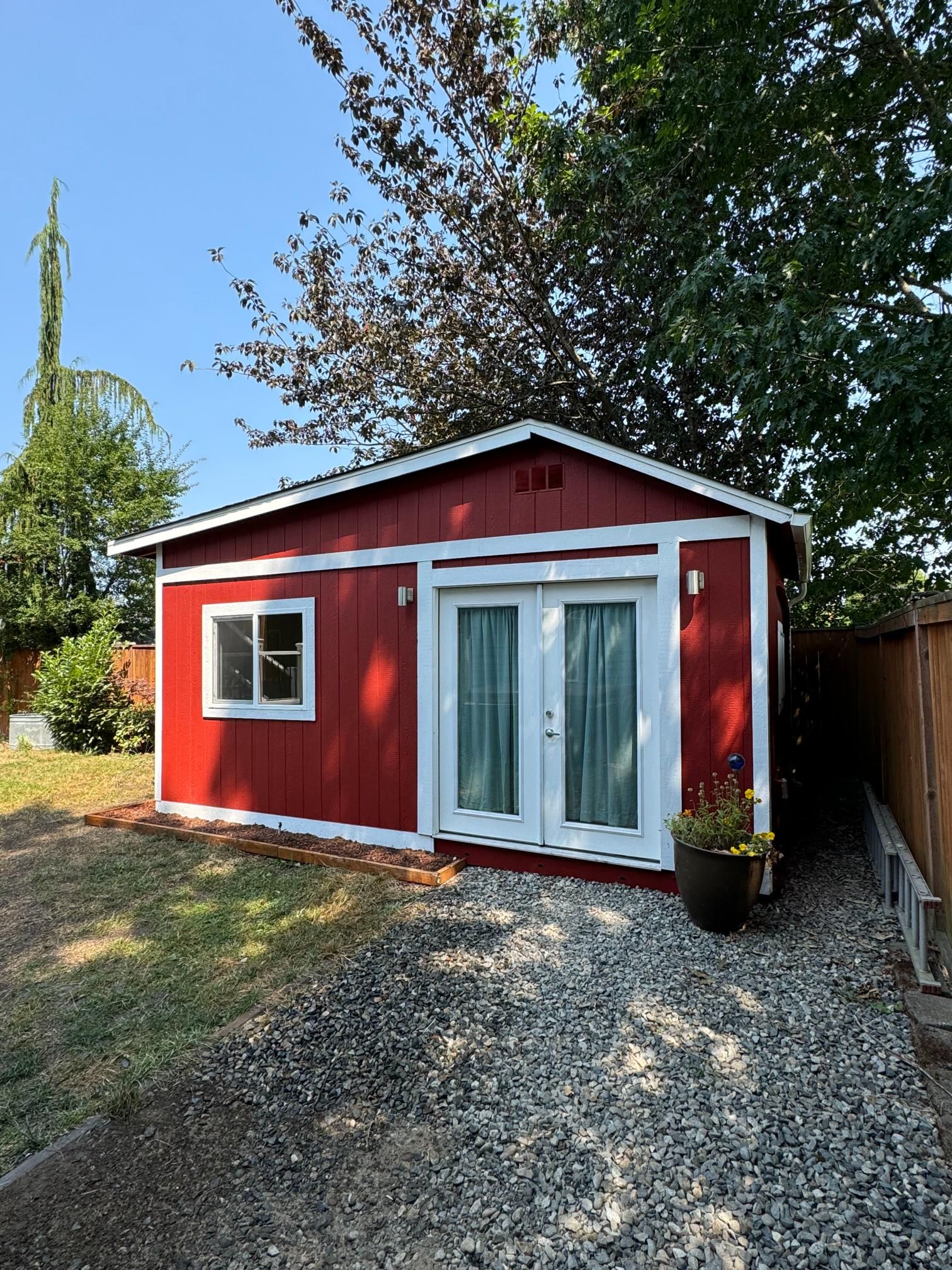 Red shed with white trim, double doors, and a window, set on gravel path, surrounded by grass and trees.
