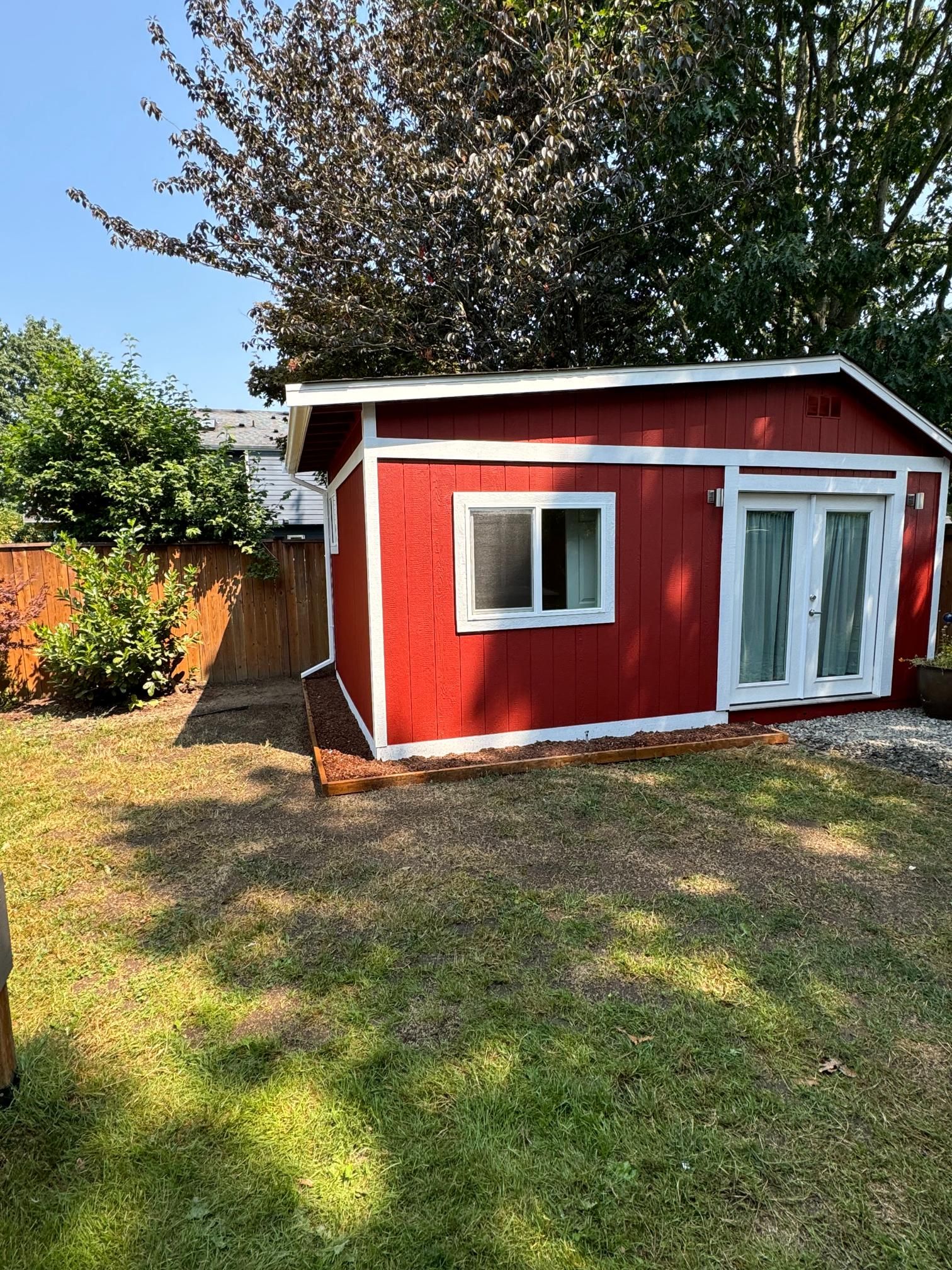 Red shed with white trim, window, and french doors in a backyard with patchy grass.