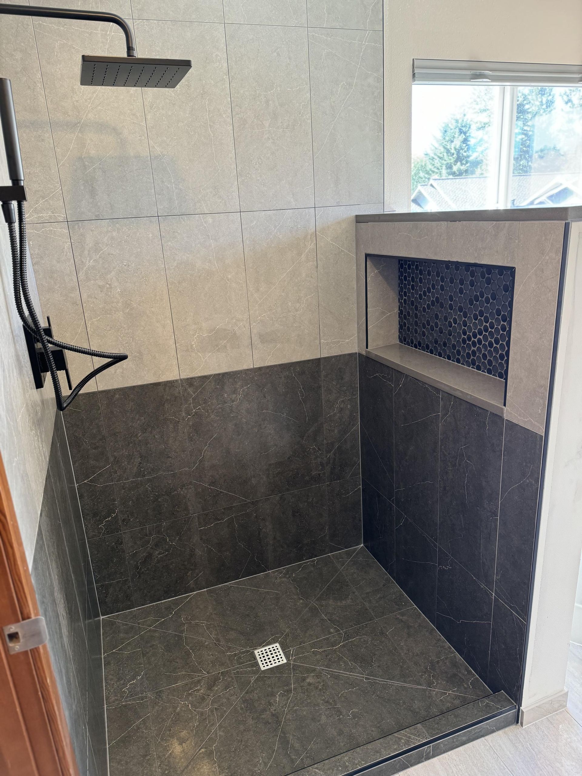 Shower with dark gray tile walls and floor, showerhead, and a built-in shelf with dark mosaic tile.