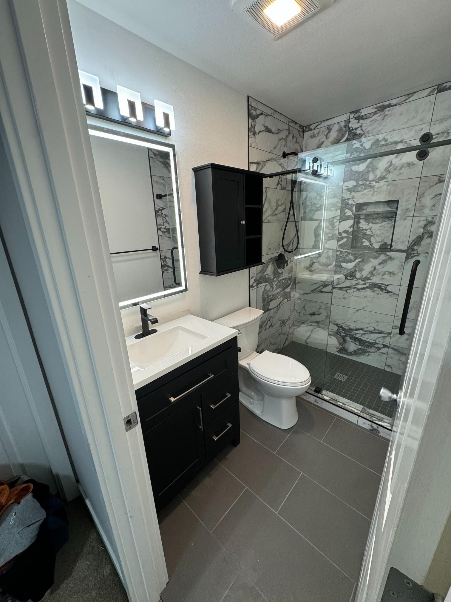Modern bathroom with black vanity, white toilet, and marble-tiled shower behind a glass door.
