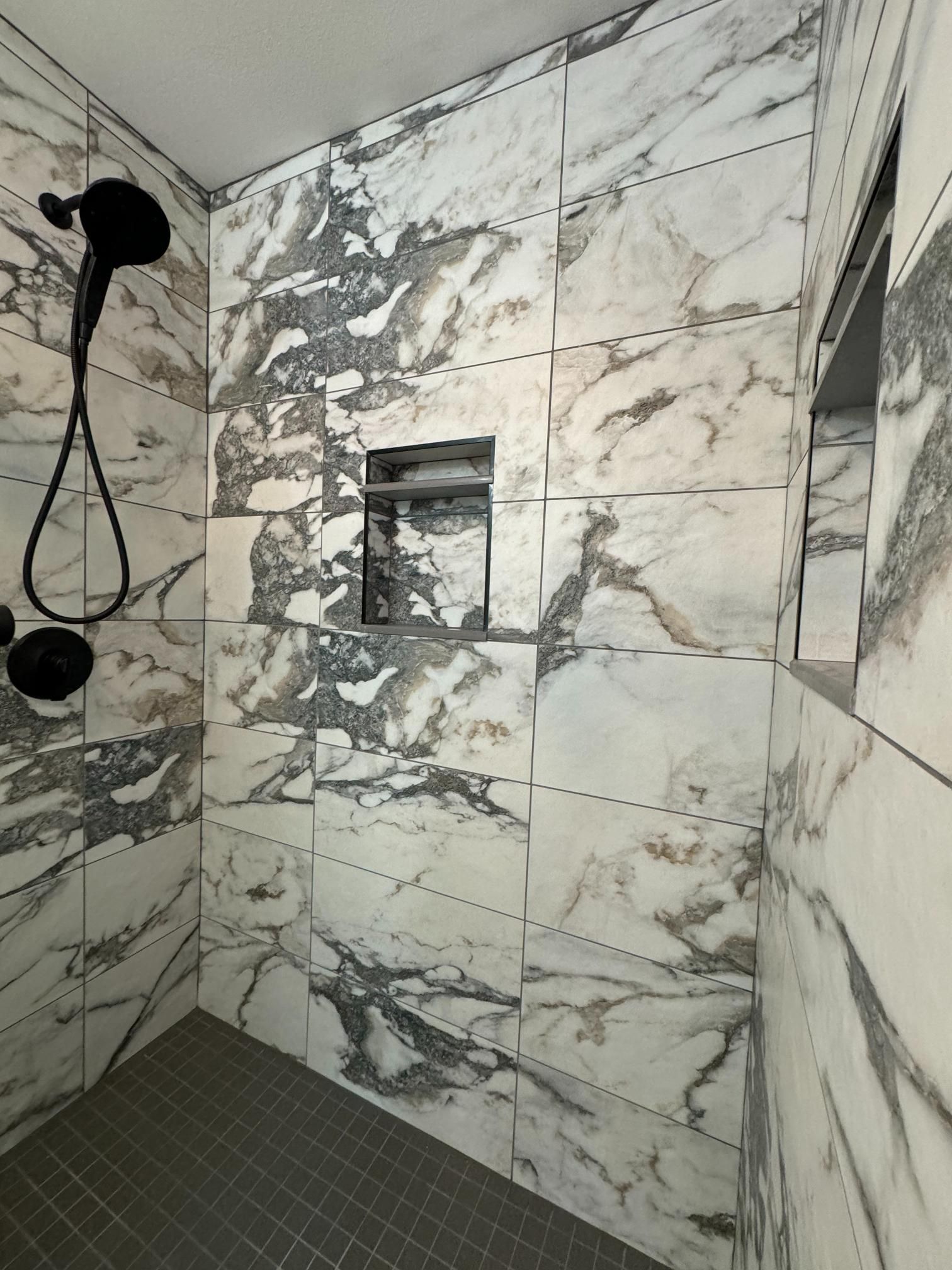 Marble-tiled shower with a black showerhead and a built-in shelf. The floor is gray.