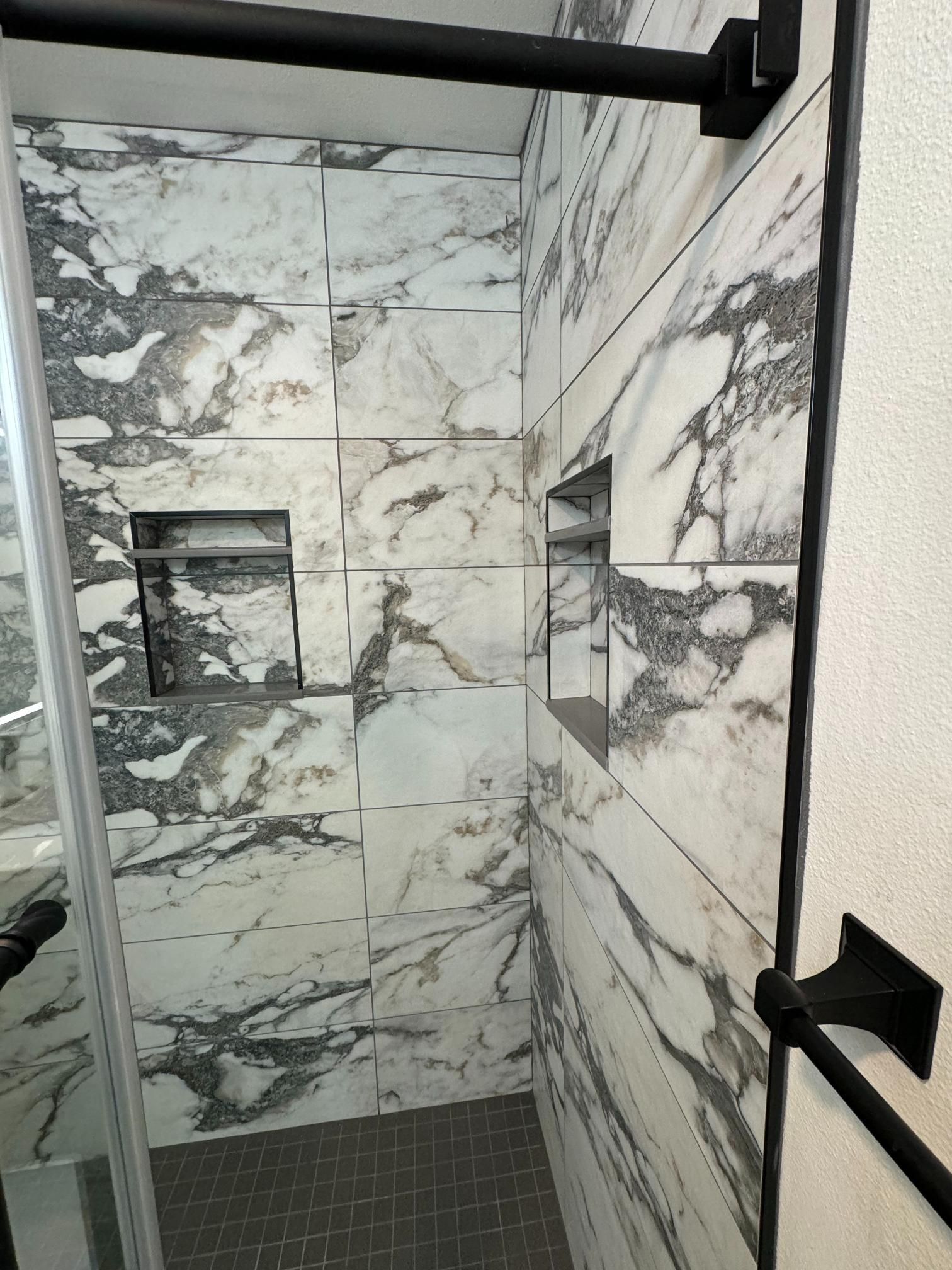 Marble-tiled shower with built-in shelves and black hardware. Grey and white patterned tiles.