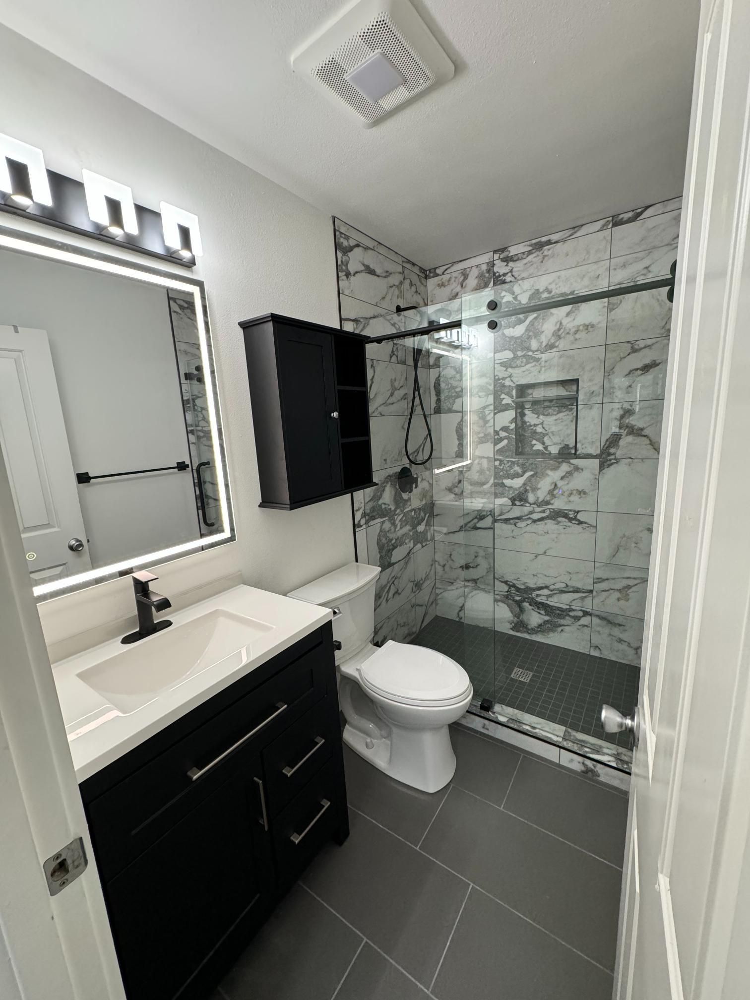 Modern bathroom with black vanity, white toilet, and marble-look shower. Gray tile flooring.