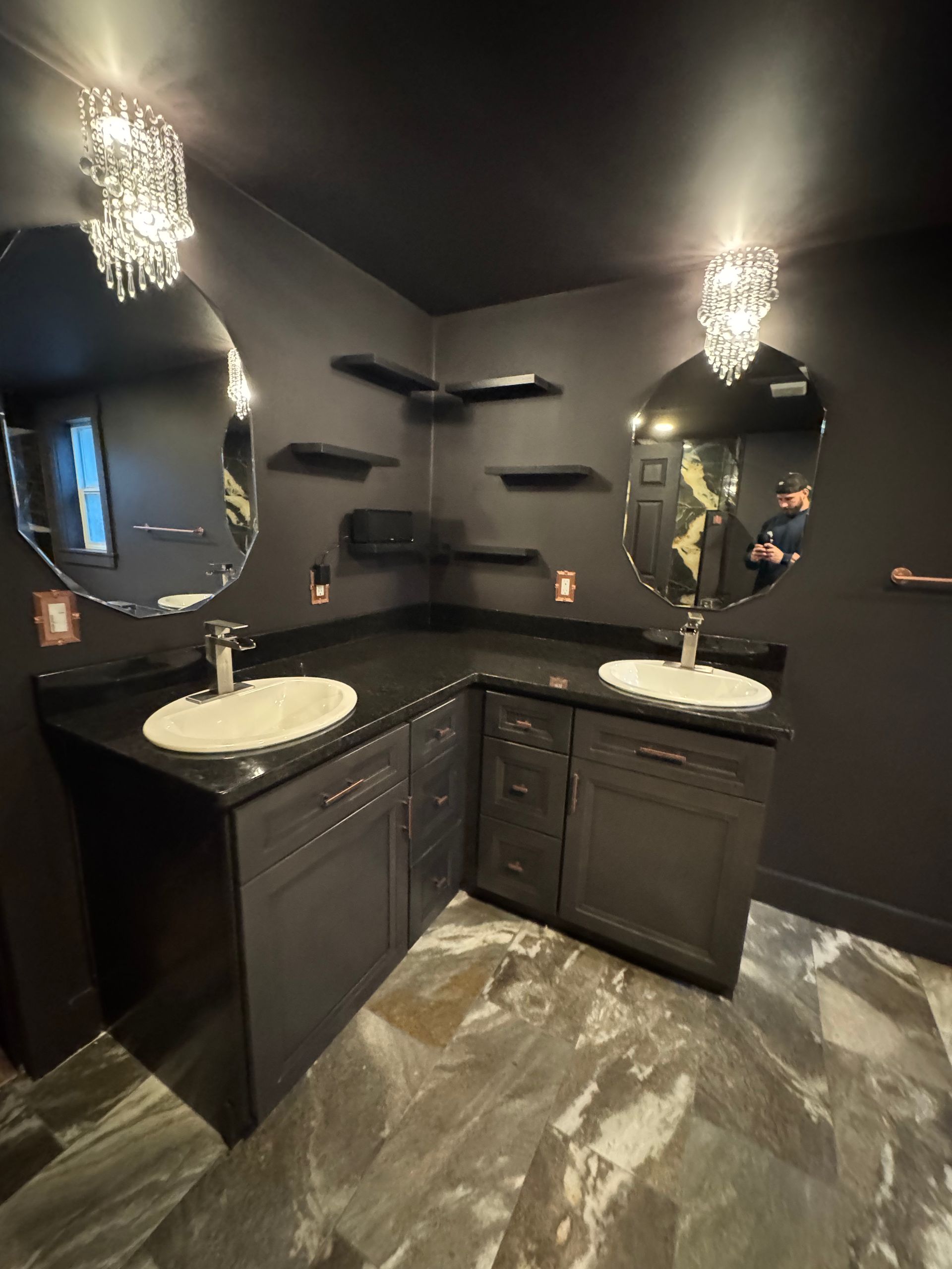 Dark gray bathroom with two sinks, oval mirrors, crystal chandeliers, and black shelves.
