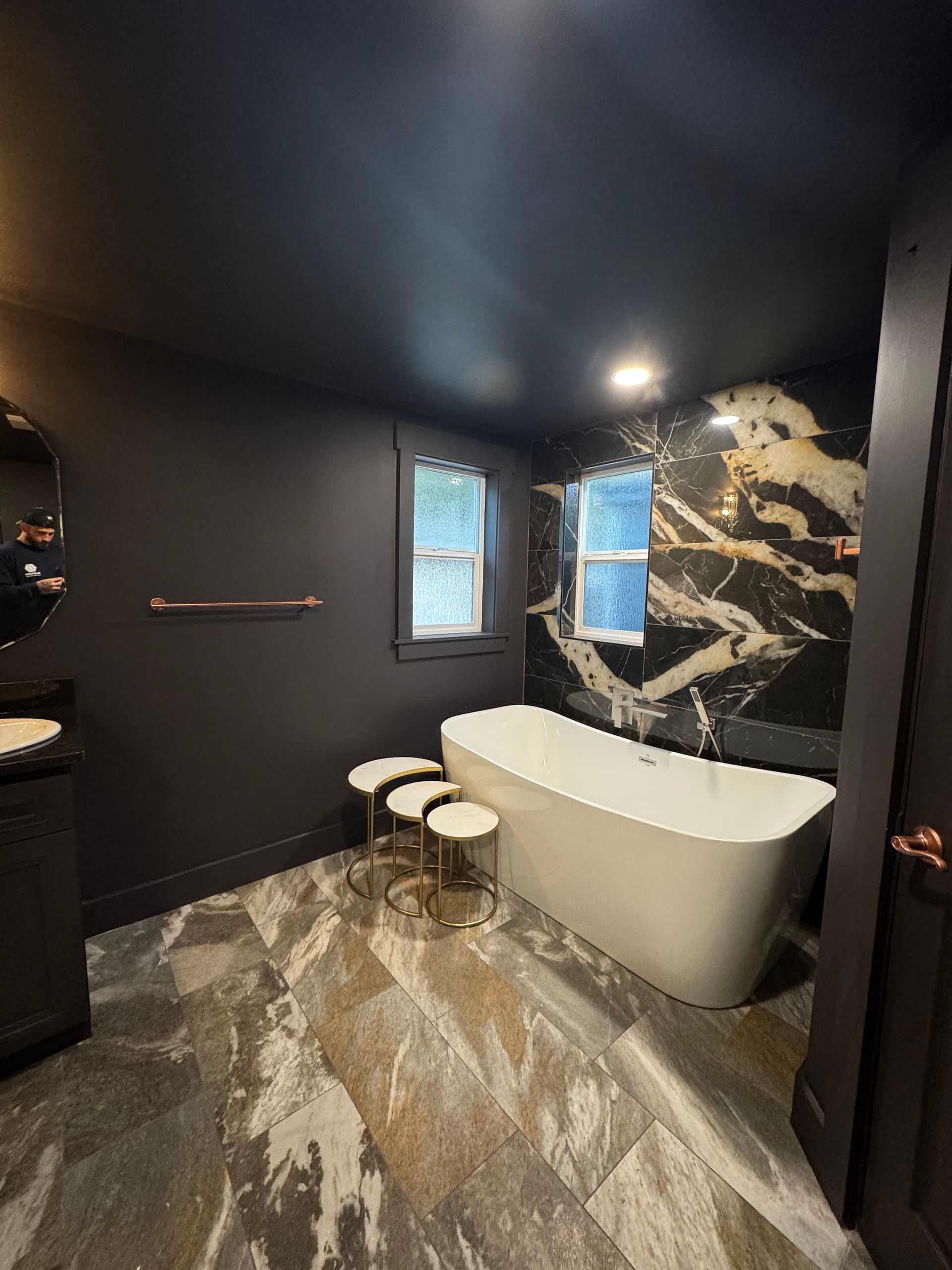 Dark bathroom with freestanding tub, black and gold marble accent wall, and patterned floor tile.