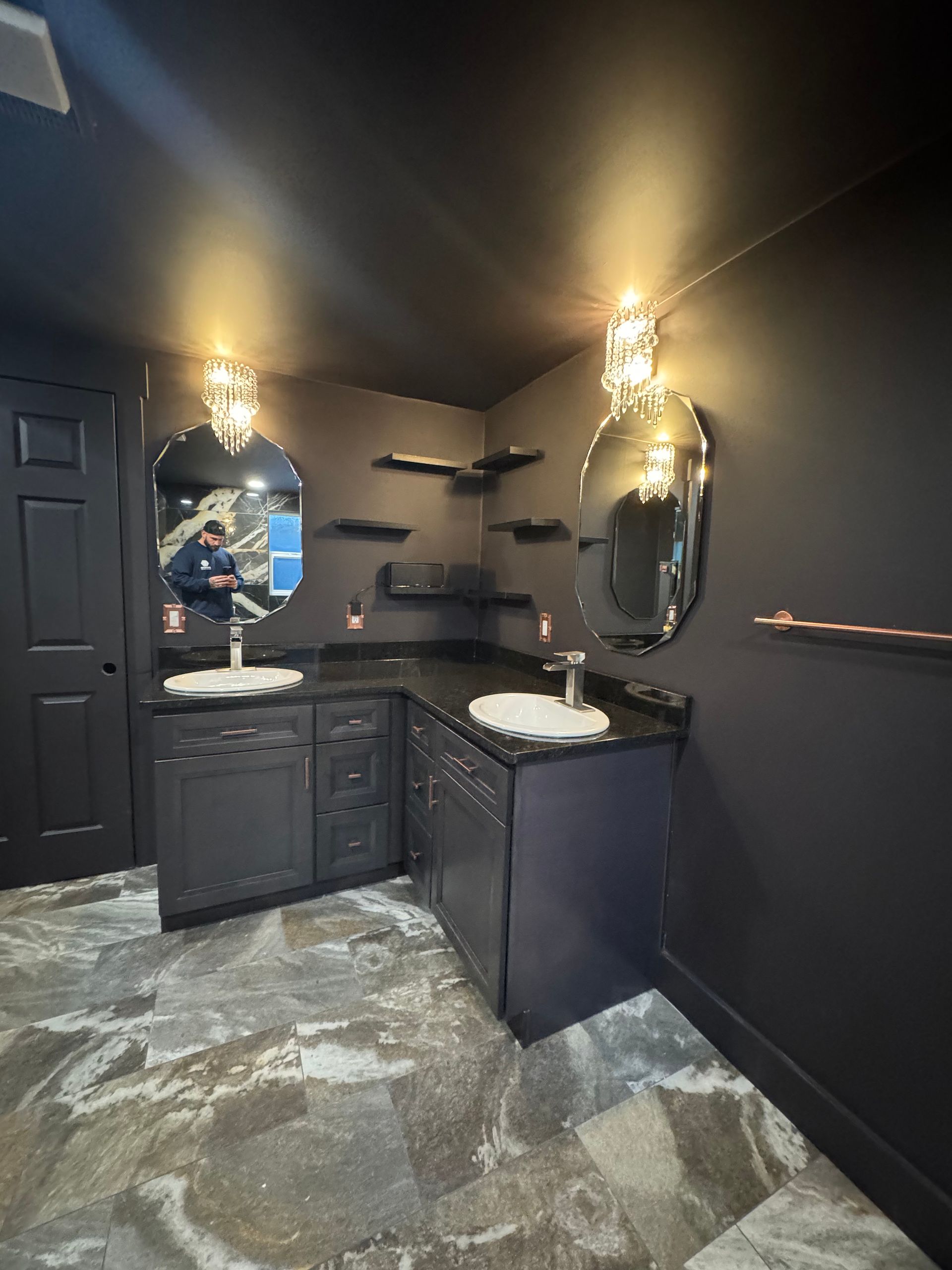 Black bathroom with two sinks, mirrors, and sconces, dark cabinetry, and patterned flooring.