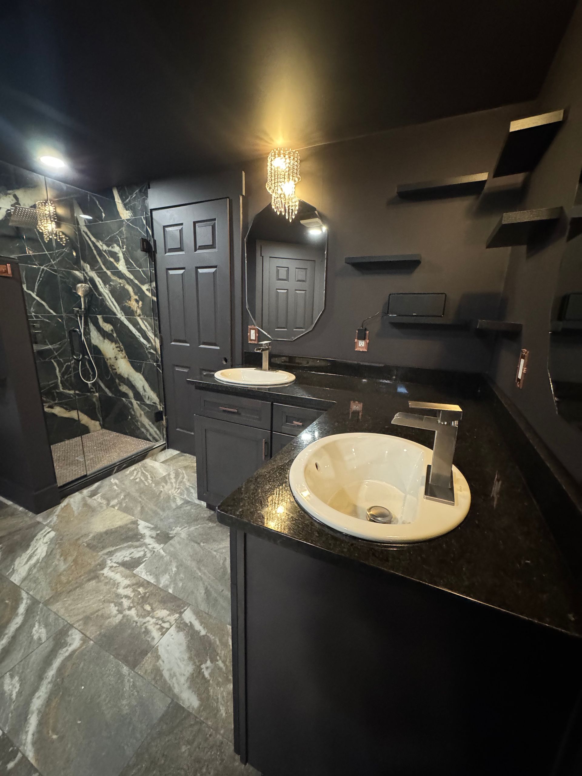 Black bathroom with granite countertops, two sinks, oval mirror, and a shower with patterned wall.
