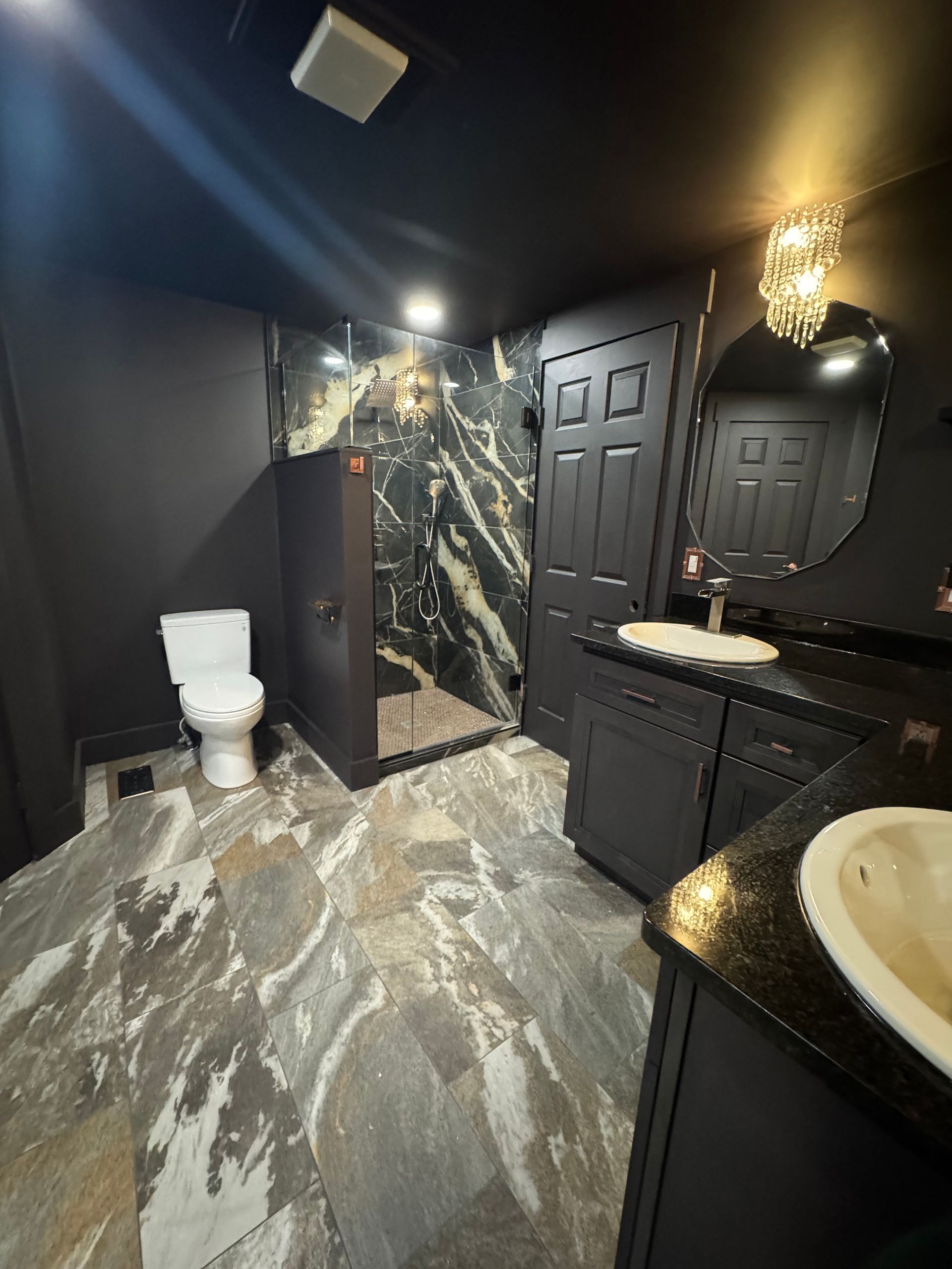 Dark bathroom with marble tile and accents. Features toilet, shower, vanity with two sinks, and black door.