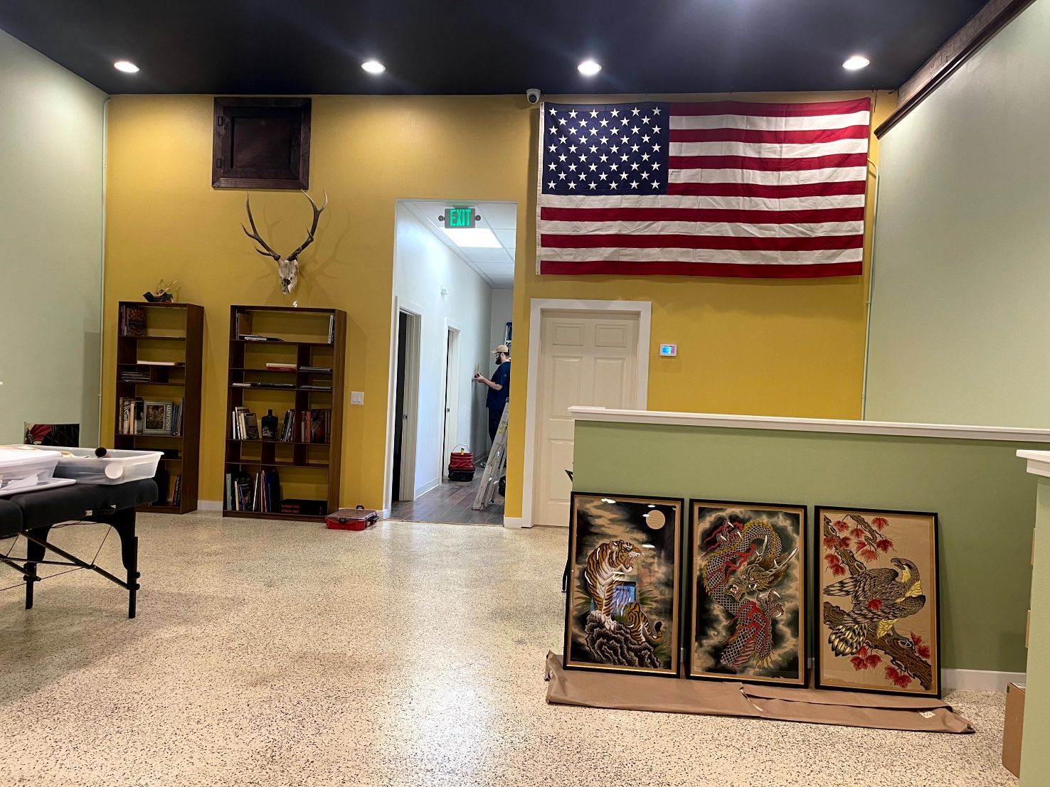 Interior room with American flag, art, shelves, yellow and green walls, and speckled floor.