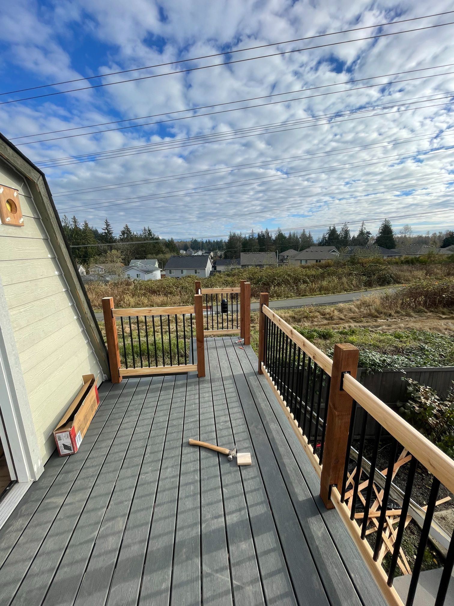 A-frame house with a composite deck and wooden railing, overlooking a field under a cloudy blue sky.