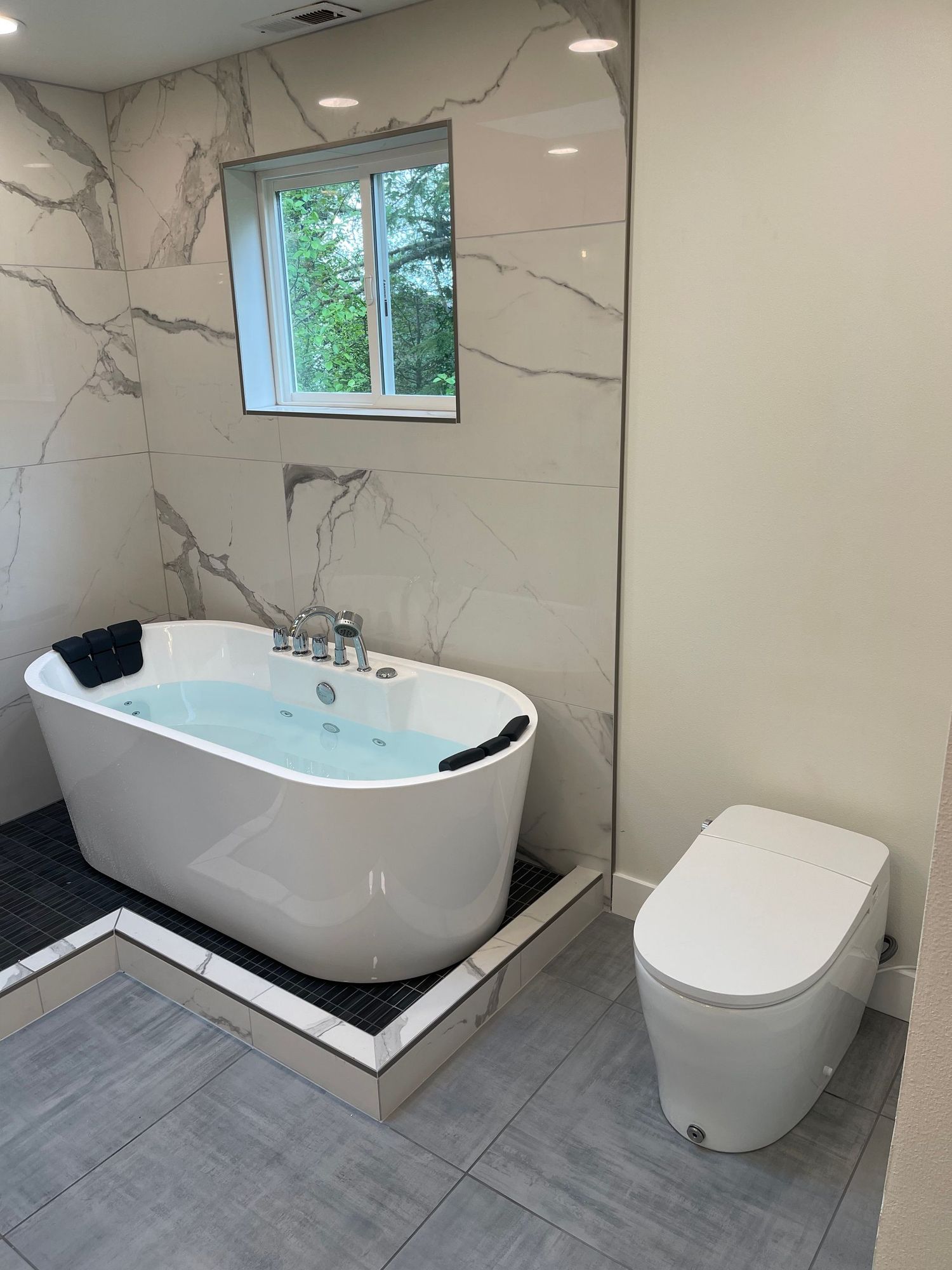 Modern bathroom with a white freestanding tub filled with water and a white toilet. Marble-look tile on the walls and flooring.