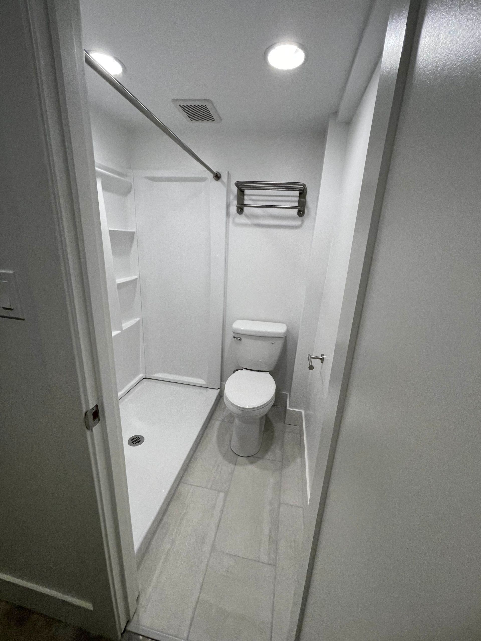 Small, white bathroom with a shower, toilet, and towel rack. Light gray flooring and recessed lighting.